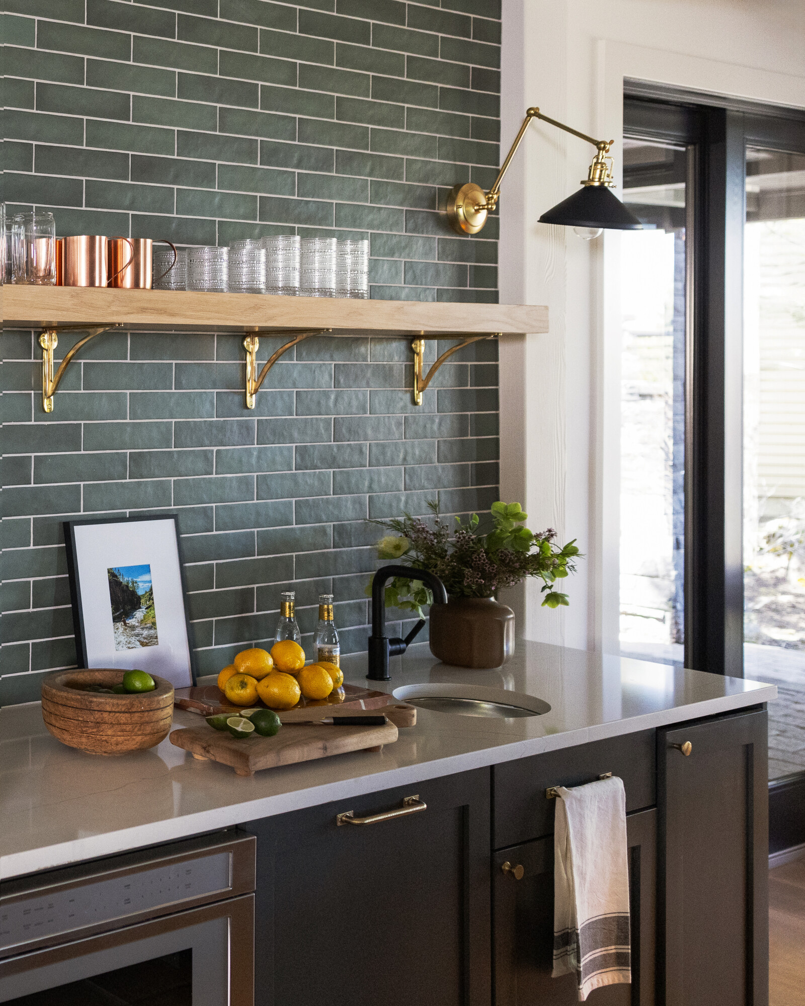 A stylish home wet bar with dark green stacked tile backsplash, a natural wood and brass open shelf holding copper mugs and glassware, a modern black and brass sconce, and a white quartz countertop. The setup includes a round sink, a vase of greenery, lemons and limes on a cutting board, and a rustic wooden bowl.