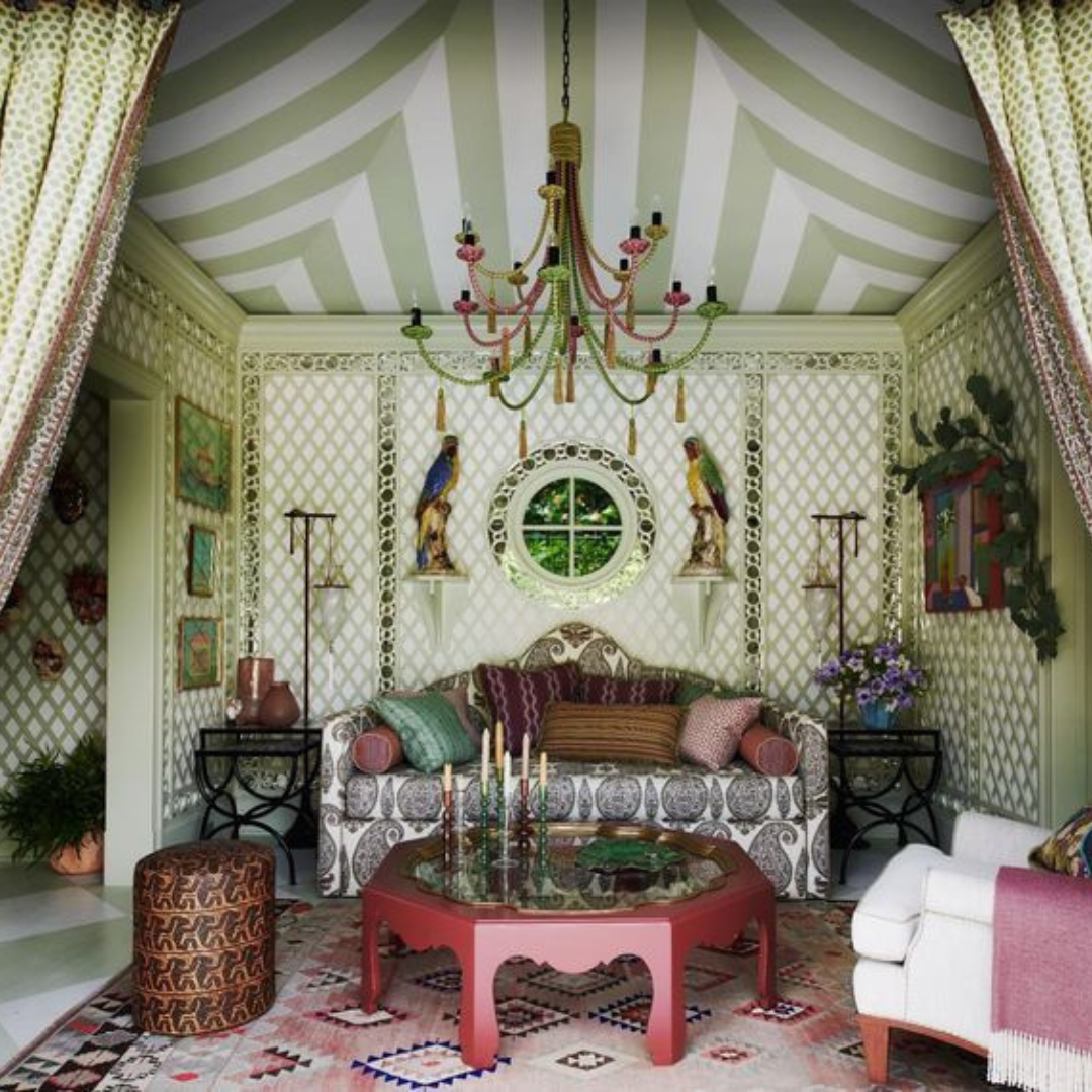 A bold and eclectic Palm Beach-style sitting room with a green and white striped tented ceiling, lattice walls, and vibrant layered decor. The space features a carved sofa with patterned upholstery, colorful pillows, a red scalloped coffee table with candles, a multicolored rug, and playful accents like parrot figurines and a beaded chandelier.