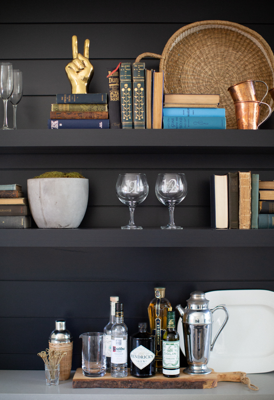 A moody and eclectic home bar setup styled on black shiplap open shelving. The shelves display a mix of vintage books, copper mugs, etched glassware, a woven basket, a gold peace sign sculpture, and a cement planter with moss. The bar surface features a wood tray with liquor bottles including Hendrick’s Gin and Dolin Vermouth, a classic silver cocktail shaker, a mixing glass, and bar tools, creating a collected, lived-in feel.