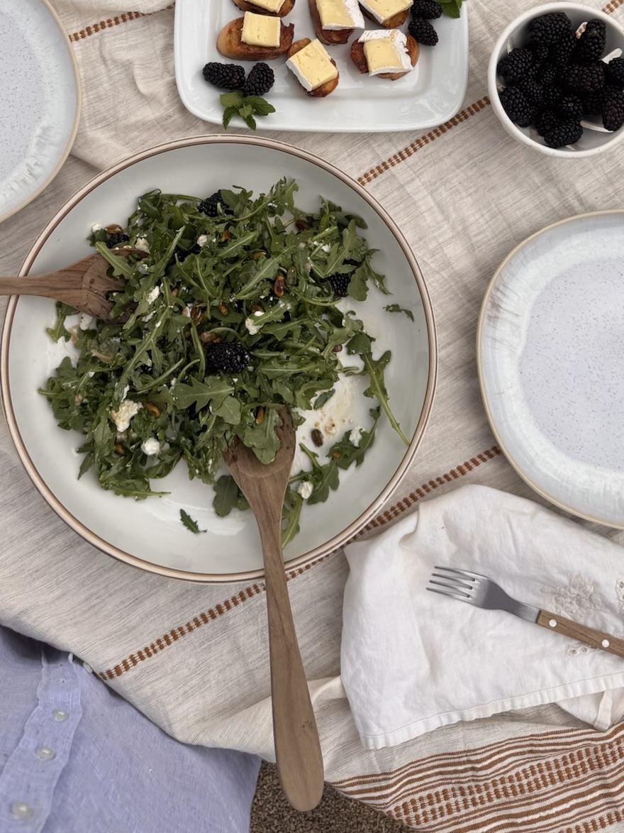 Overhead view of a summer picnic setup featuring a large arugula salad with blackberries, goat cheese, and toasted nuts in a wide ceramic bowl with wooden servers. The table is set with neutral-toned dishes, a small bowl of fresh blackberries, and a plate of maple-brushed baguette slices topped with brie. Everything rests on a soft striped linen tablecloth, creating a relaxed and inviting outdoor dining moment.