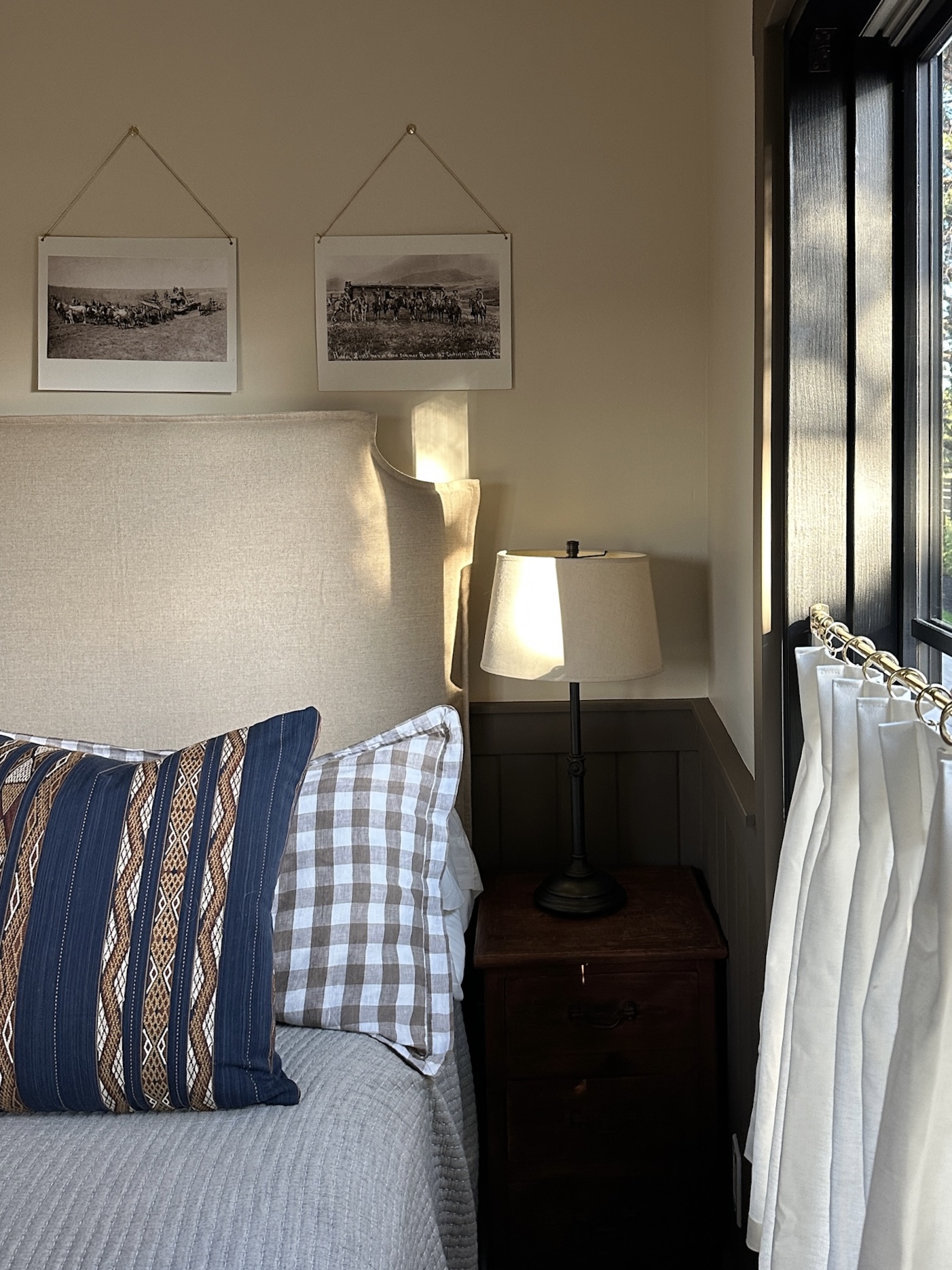 Neutral bedroom scene with dark olive wainscoting, soft pleated cafe curtains filtering natural light, and a glimpse of surrounding trees through the window screen.