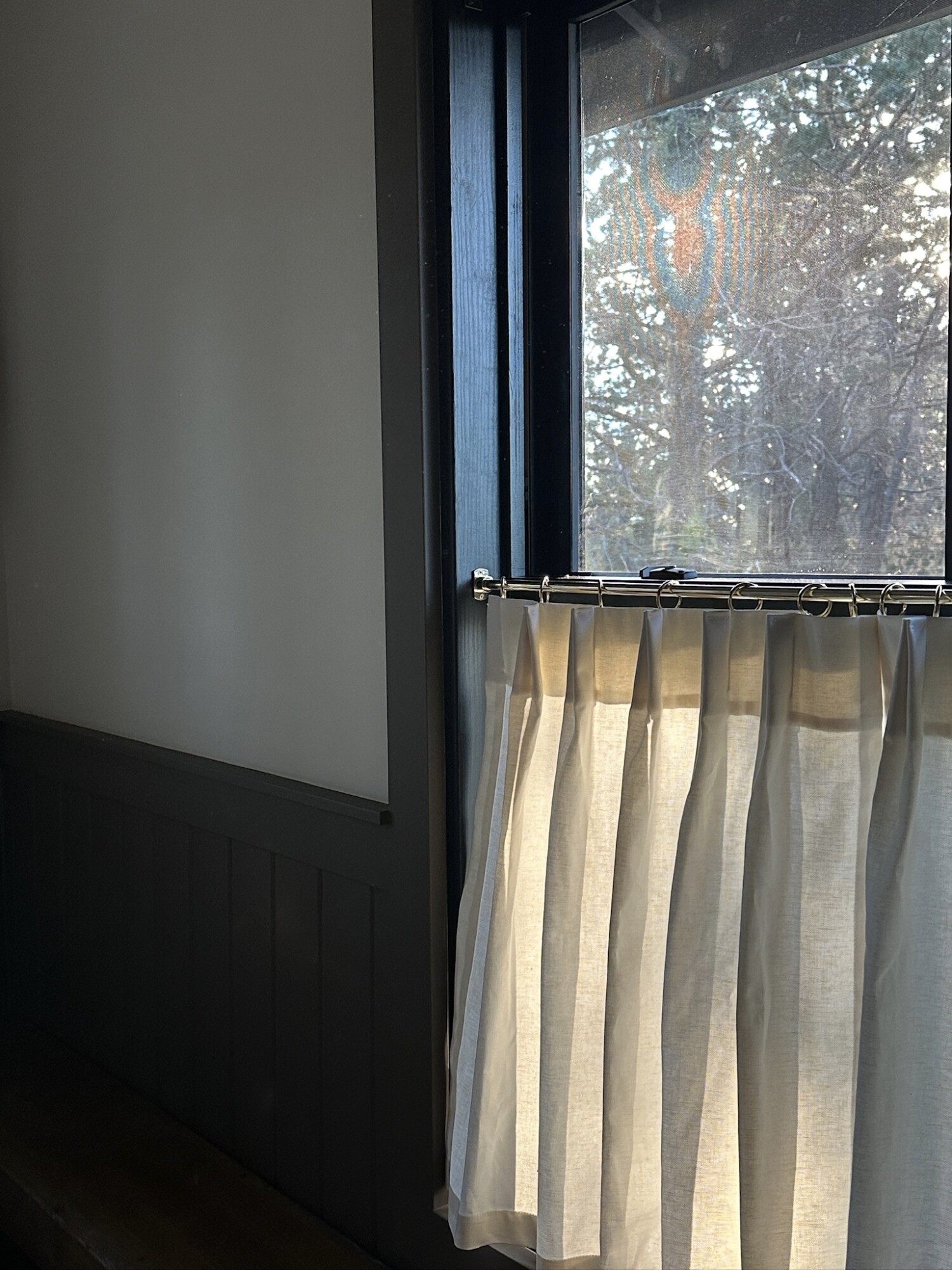 Close-up of a black-trimmed window with crisp white cafe curtains hung on a chrome rod, softly lit by late afternoon sunlight streaming in.