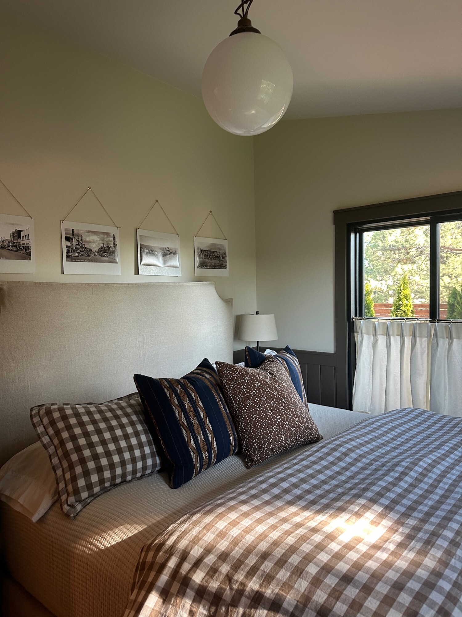 Cozy guest bedroom corner with a light upholstered headboard, layered brown gingham and vintage-patterned pillows, dark wood nightstand with a lamp, and soft cafe curtains partially covering a black-framed window.