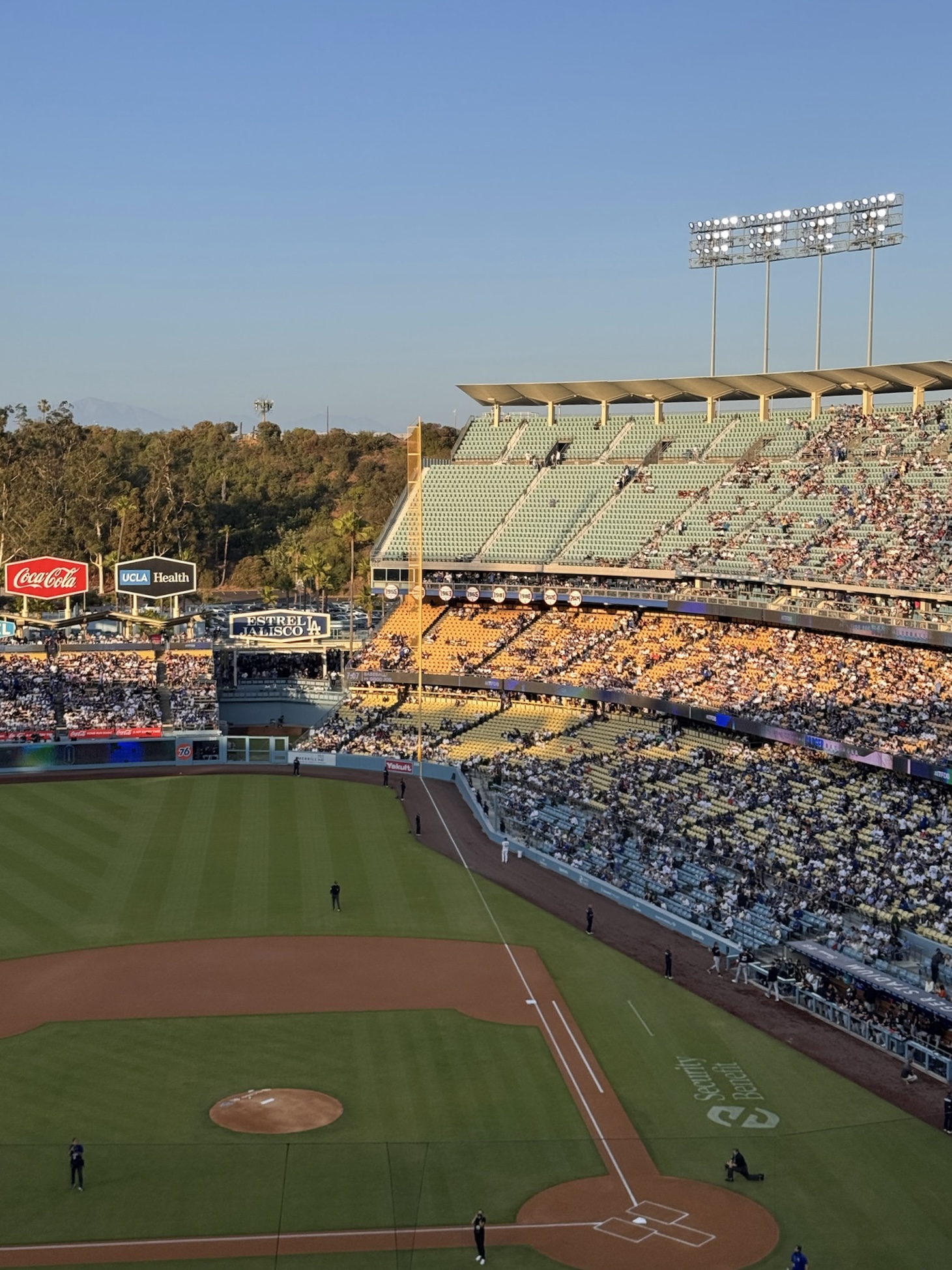 View of Dodger Stadium at sunset with the baseball field in the foreground and the stands partially filled with fans, golden hour light casting warm tones over the crowd.