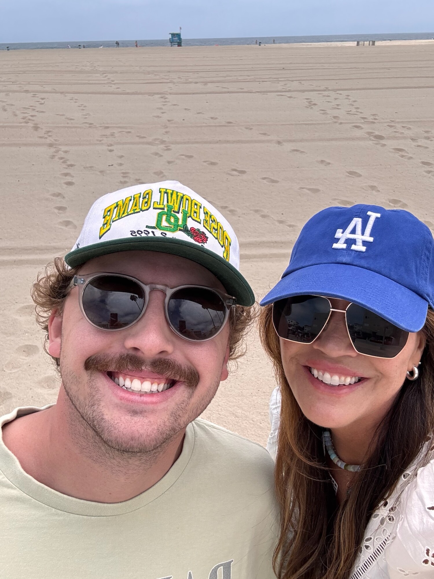 Smiling couple taking a selfie on a wide, empty beach with soft sand and a hazy blue sky in the background. The woman wears a blue LA Dodgers cap and sunglasses; the man wears a vintage Rose Bowl Game cap and sunglasses.