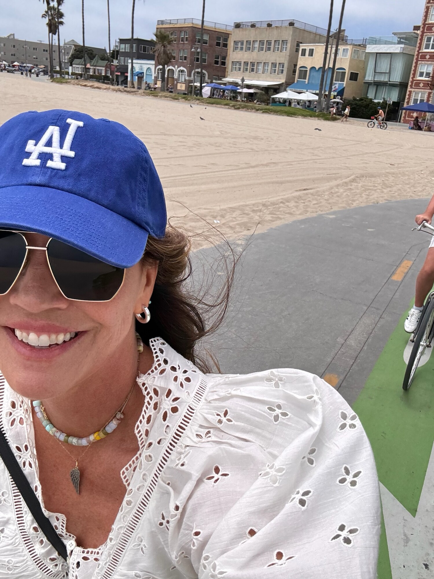 Woman smiling while riding a bike along the beach boardwalk in Santa Monica, wearing a blue Dodgers baseball cap, sunglasses, and an embroidered white eyelet blouse.