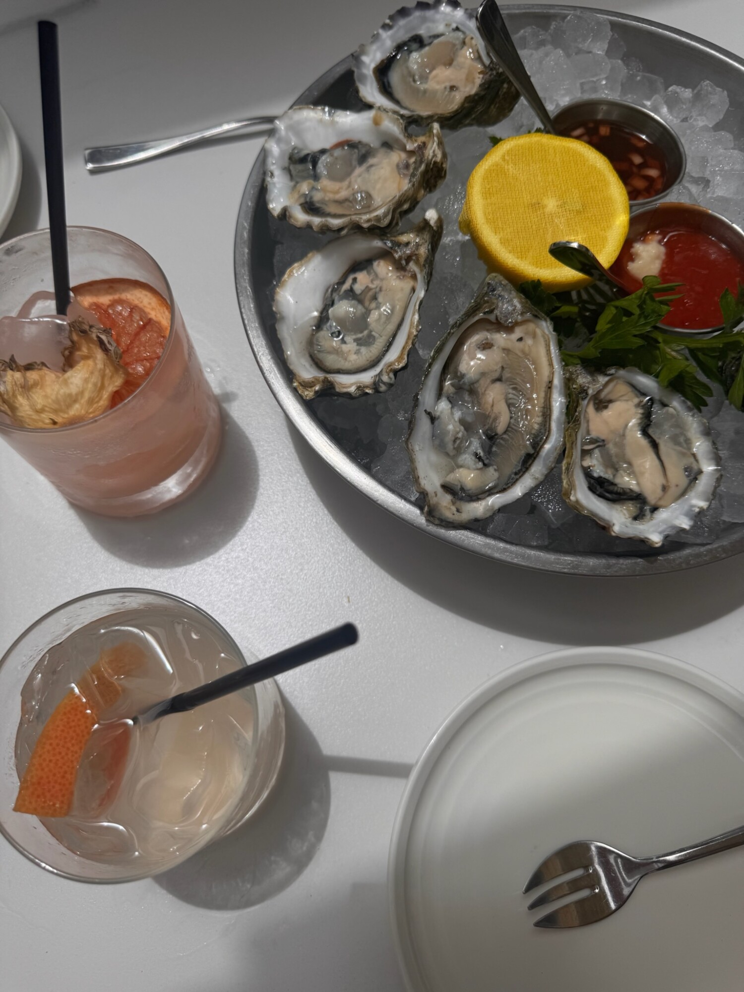 Overhead shot of a seafood platter with fresh oysters on ice, cocktail sauces, and a lemon slice, surrounded by two grapefruit cocktails and small plates. 