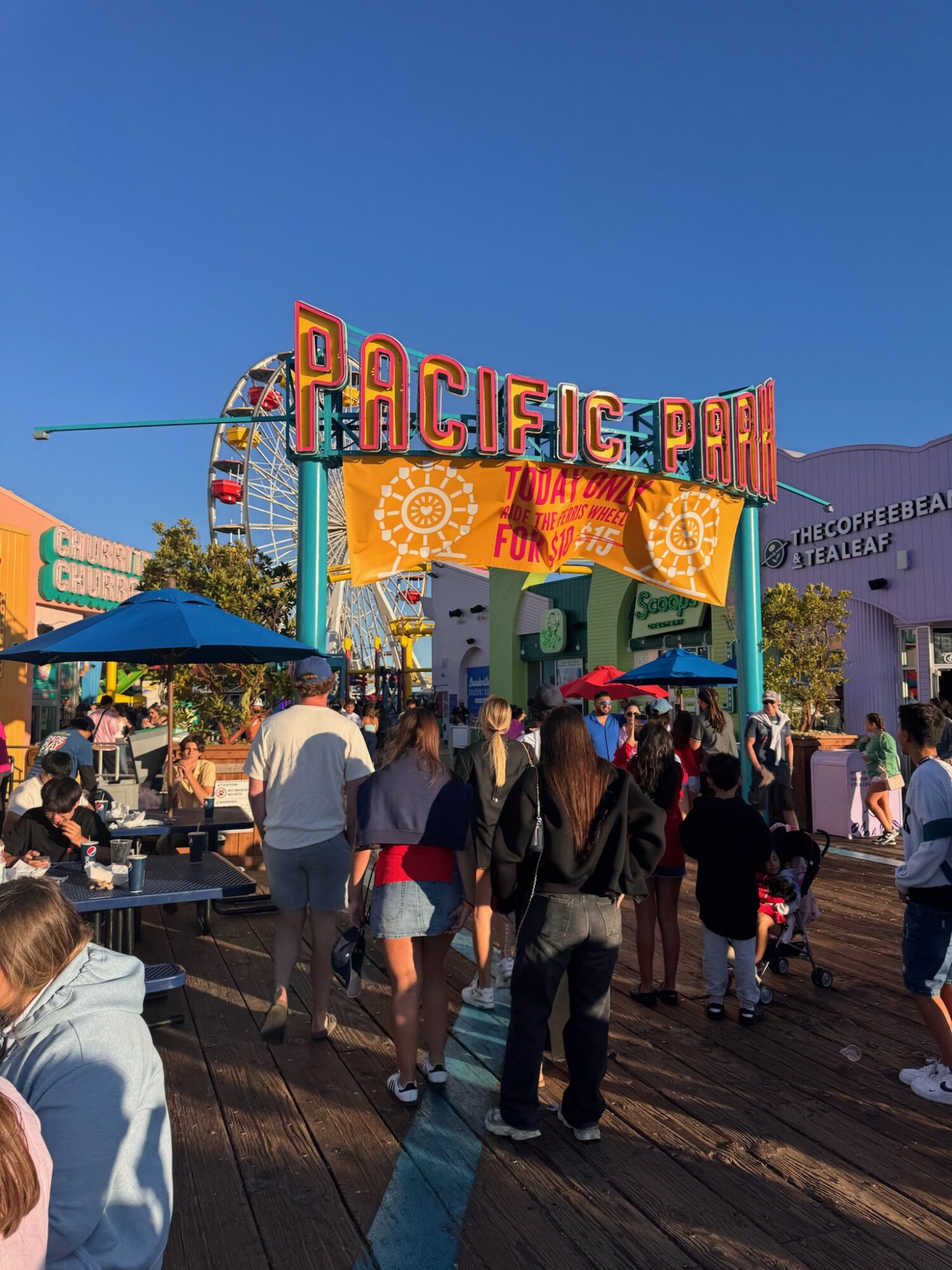 Crowds walking toward the colorful entrance of Pacific Park at the Santa Monica Pier, with the iconic Ferris wheel and bright signage under a clear blue sky.