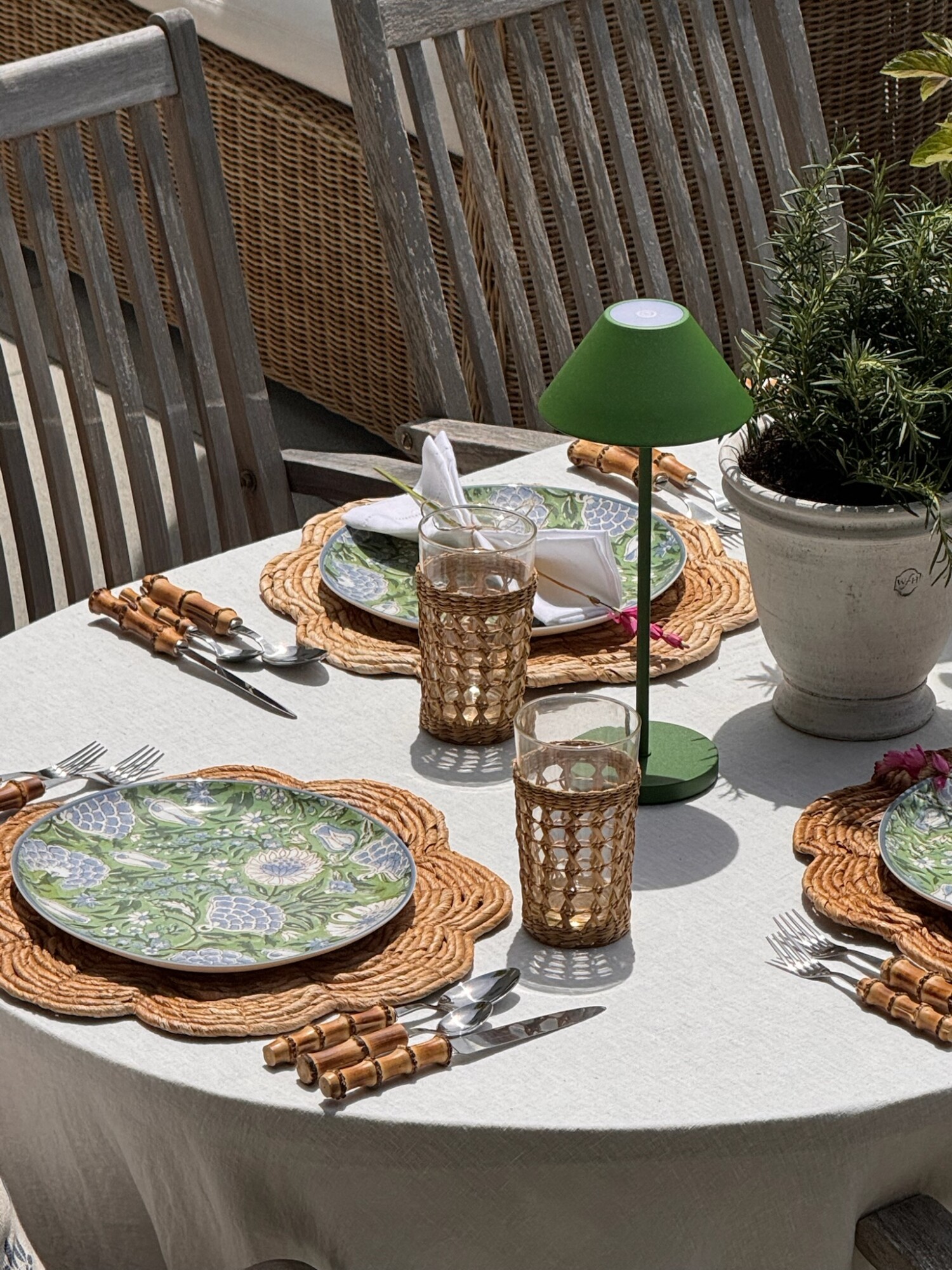 An outdoor summer dining table set with woven placemats, green and blue patterned plates, bamboo-handled flatware, and vintage-inspired amber glasses. A green rechargeable table lamp and a white planter with fresh rosemary add a charming and functional centerpiece to the natural, sunlit setup.