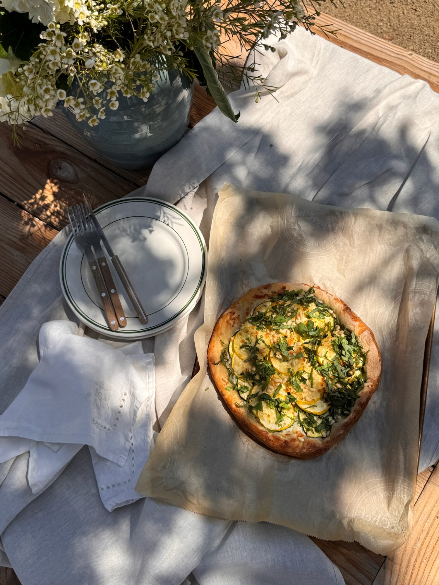 A relaxed outdoor table setup with fresh herbs, white floral arrangement, and a golden-baked squash galette catching warm afternoon light.