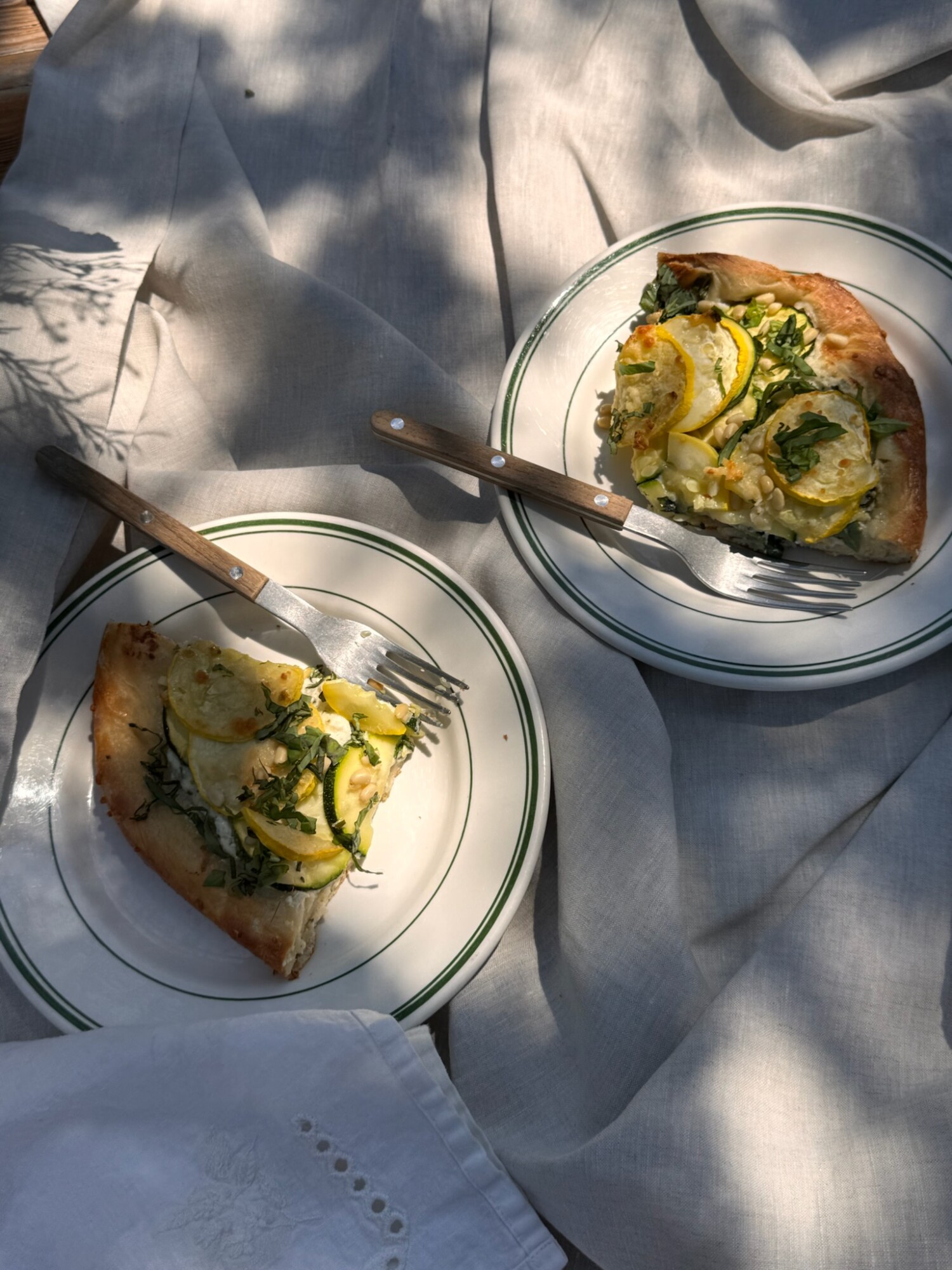 Two slices of squash galette are served on white-rimmed vintage plates with wooden-handled forks, resting on soft linen fabric in dappled sunlight.