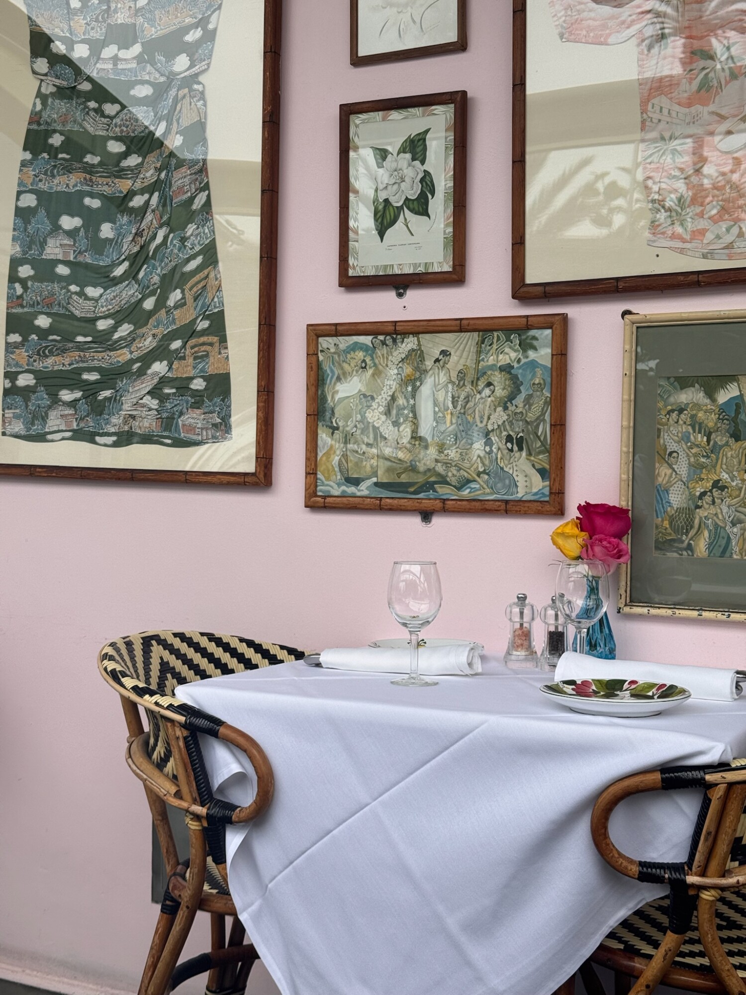 A small outdoor dining table set for two with a white tablecloth, rattan bistro chairs, and a glass vase of fresh roses, set against a pink wall adorned with framed Japanese art and floral illustrations.