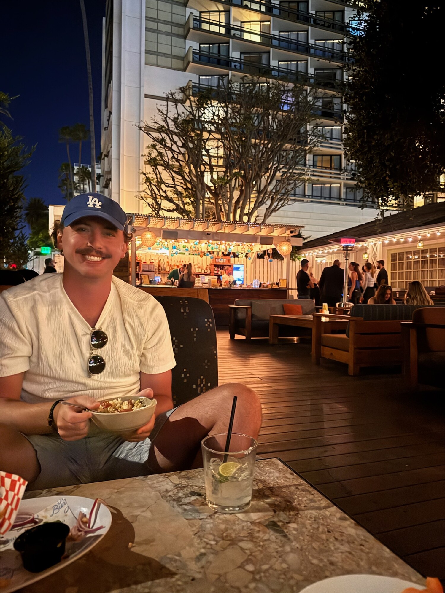 Man in a white shirt and blue Dodgers cap smiling while enjoying a bowl of food at an outdoor patio restaurant in Santa Monica at night, with festive lights and people dining in the background.