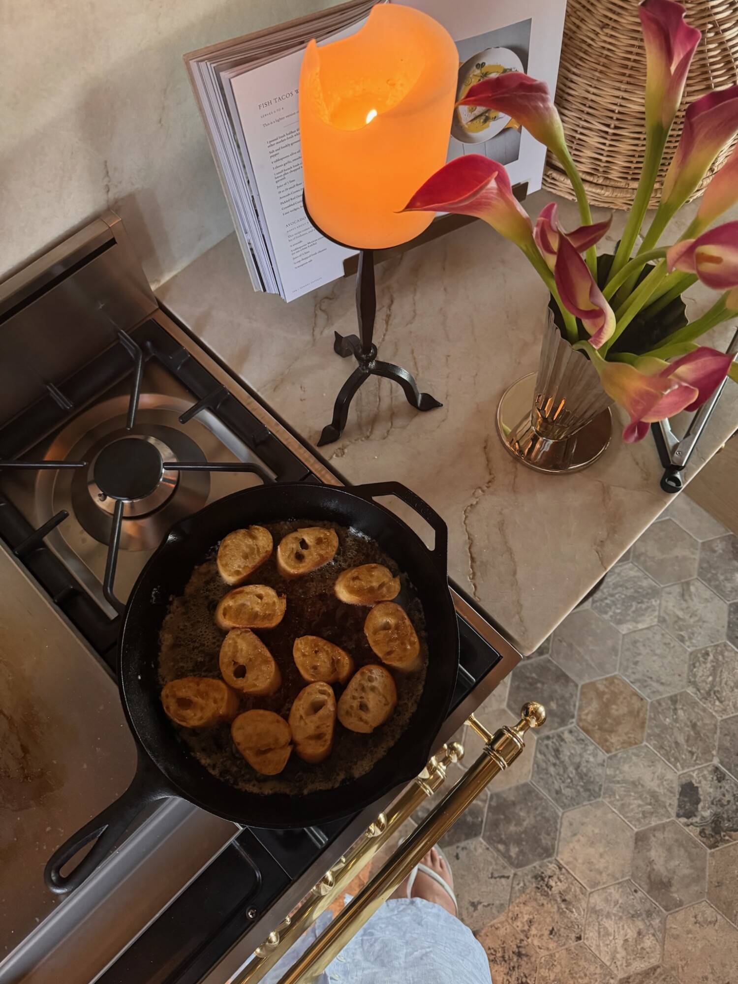 Cozy kitchen scene with a cast iron skillet toasting baguette slices in butter on a gas stove. A glowing candle, cookbook, and vase of pink calla lilies sit on the marble countertop, with warm-toned hexagon tile flooring visible below. A relaxed, ambient moment captured mid-recipe.