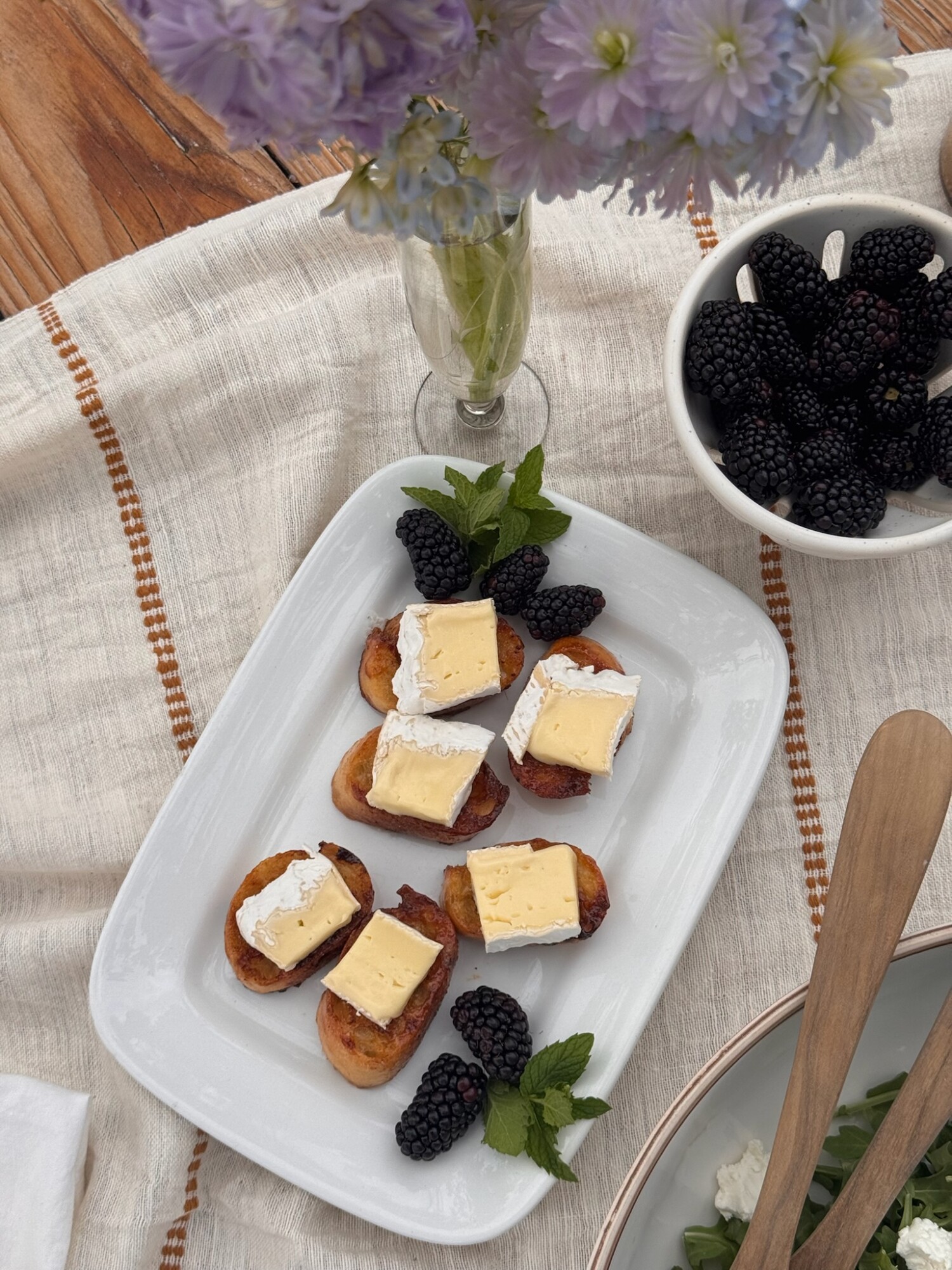 Overhead view of a summer appetizer spread featuring toasted baguette slices topped with brie and maple glaze, garnished with fresh blackberries and mint on a white platter. A bowl of blackberries, a wooden salad bowl, and a vase of light purple flowers sit nearby on a striped linen tablecloth.
Let me know if you want a Pinterest pin caption or story text to go with it!