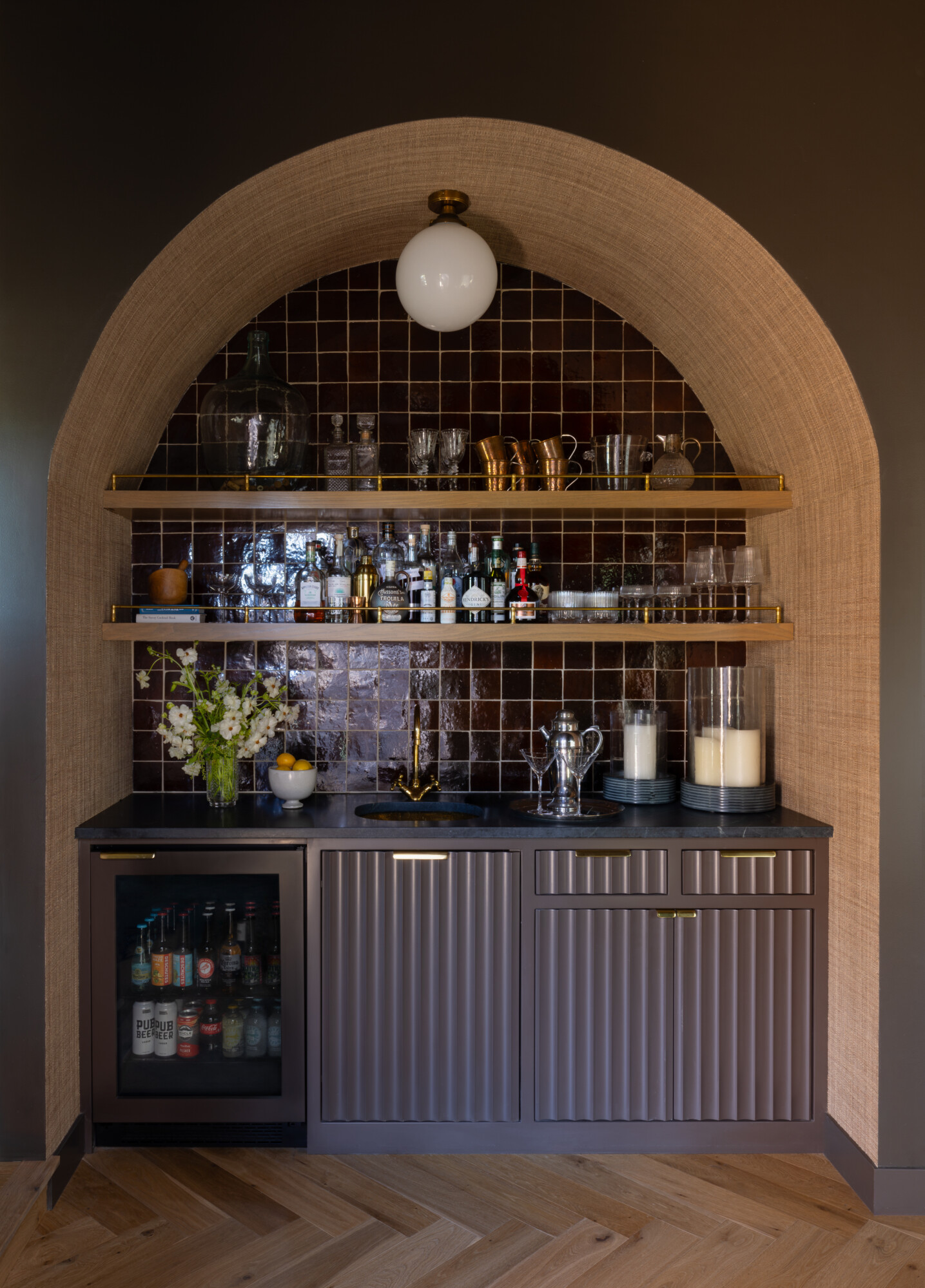 A moody and elegant wet bar built into an arched alcove. The space features dark, glossy square tile backsplash, grasscloth-wrapped archway, and ribbed cabinetry in a warm mauve-gray tone. Open wood and brass shelving displays liquor bottles, glassware, and bar accessories. A compact black sink and gold faucet sit beneath a modern globe sconce.