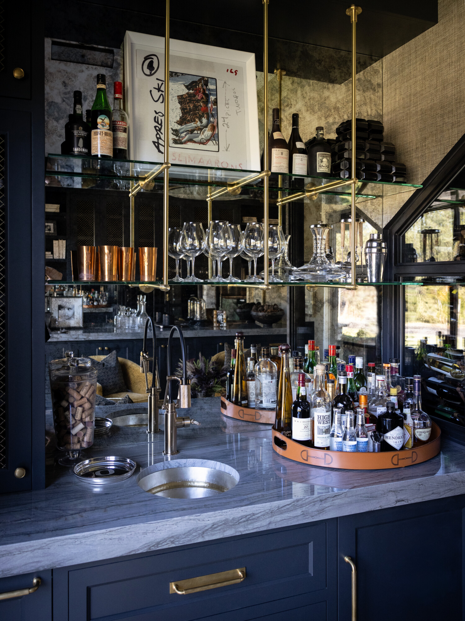 A sophisticated wet bar with dark navy cabinetry, brass hardware, and mirrored shelving. Features include a small round sink, polished marble countertops, brass faucet, and two leather-wrapped trays stocked with assorted liquor bottles. Glassware and copper cups are displayed on floating glass shelves with brass supports. A framed Apres Ski print leans casually on the upper shelf.