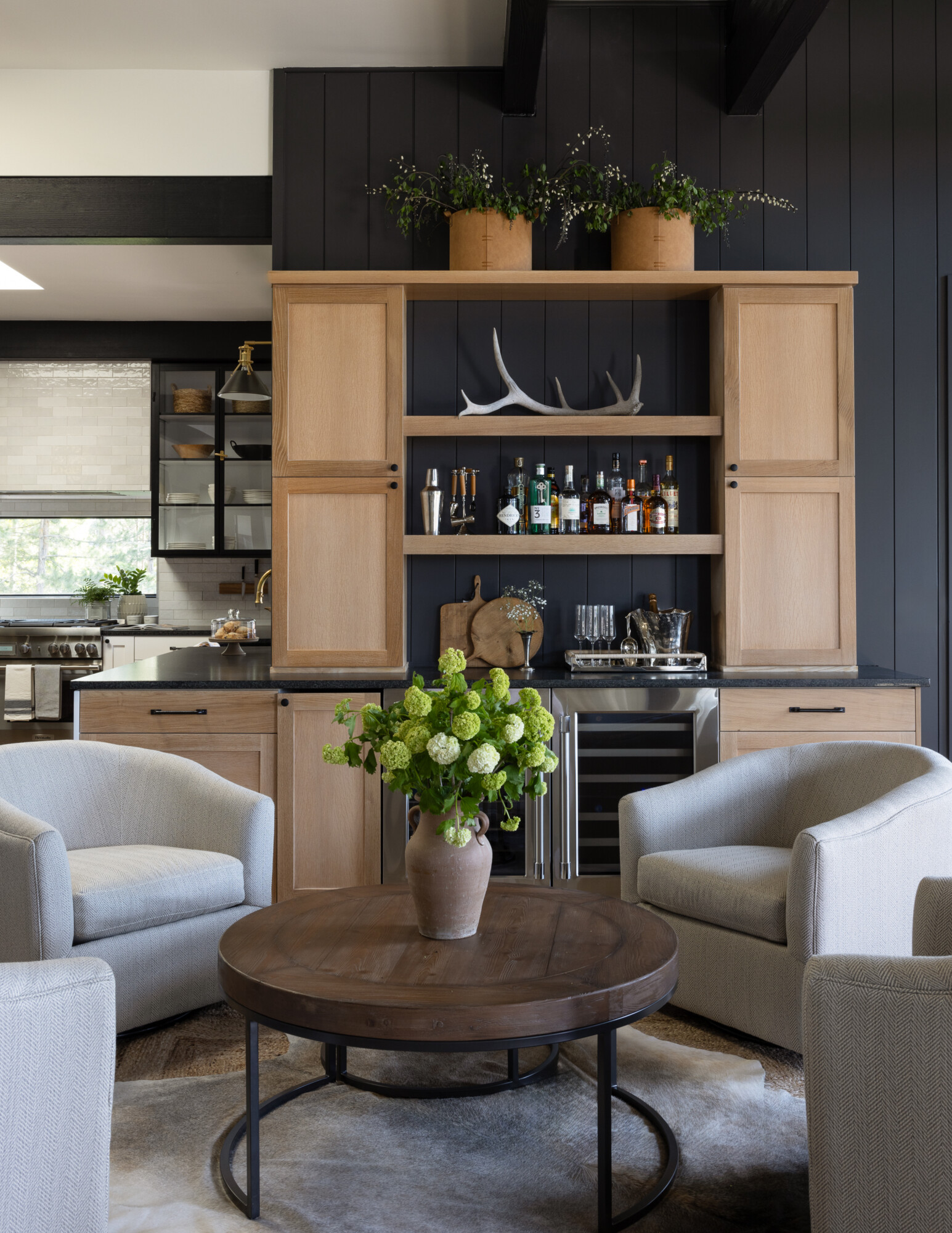 Stylish home bar setup with built-in light wood cabinetry against a dark vertical shiplap wall. Open shelving displays liquor bottles, cocktail tools, and natural accents like antlers and wood cutting boards. Three terracotta planters with greenery line the top shelf. In front, a round dark wood coffee table with a rustic terracotta vase filled with green hydrangeas is surrounded by four textured cream swivel chairs. The space feels cozy, refined, and perfectly layered with soft lighting and contrast.