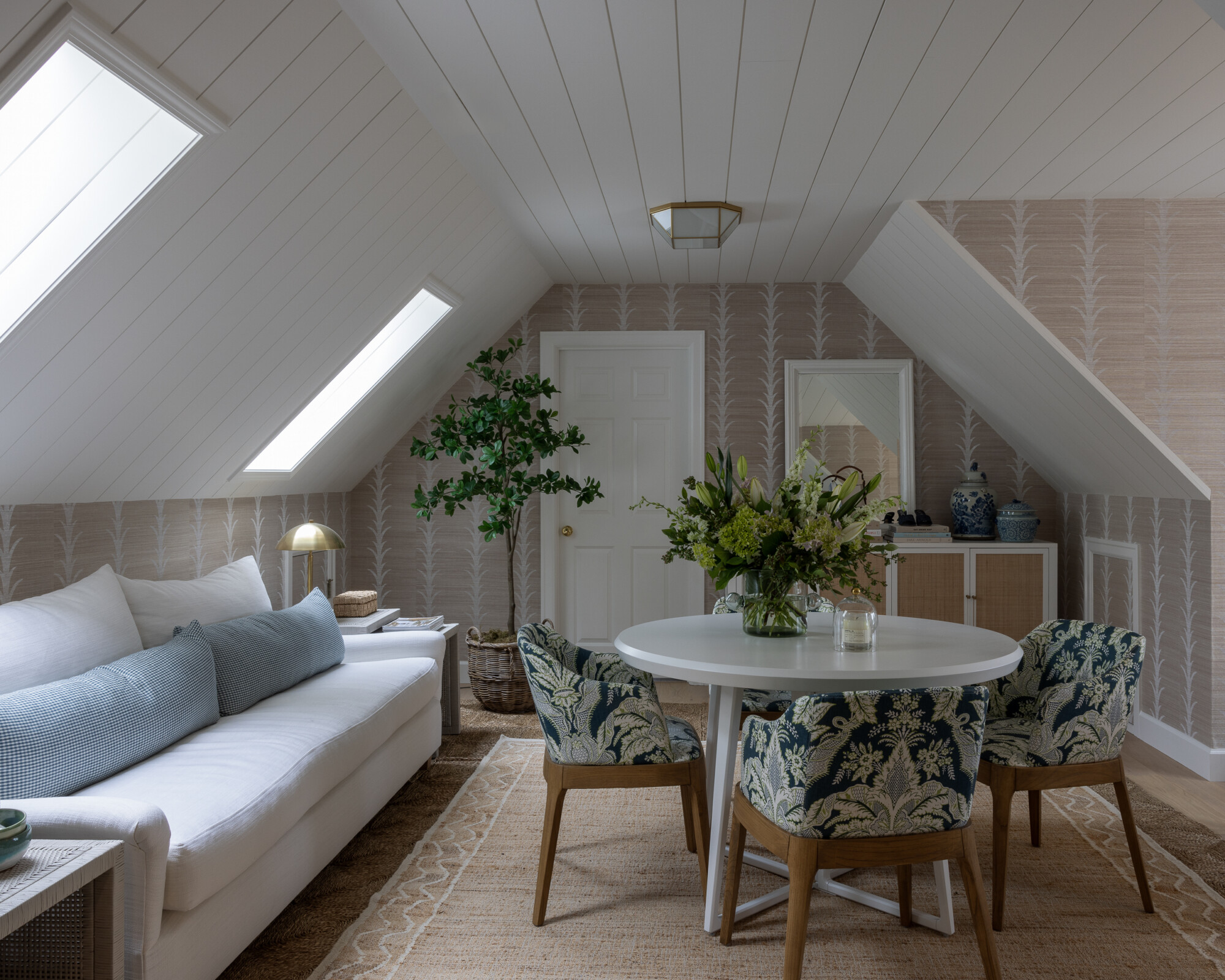 Charming attic room with sloped ceilings, painted white tongue-and-groove paneling, and skylights that flood the space with natural light. A long, white slipcovered sofa sits on a woven jute rug, accented with soft blue pillows and a brass mushroom lamp. A white round pedestal table is surrounded by four floral-upholstered dining chairs with wooden legs. In the back, a console with cane-front doors is topped with blue-and-white ginger jars and a mirror. Soft wallpaper with a subtle botanical motif adds warmth and texture throughout the room, creating a cozy, elevated retreat.