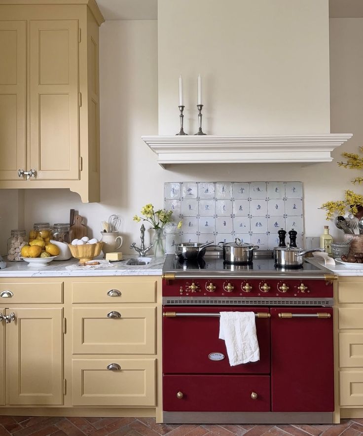 A charming cottage kitchen with buttery yellow cabinetry, a burgundy La Cornue range, and Delft-style tiled backsplash. The counter is styled with lemons, eggs, and vases of yellow flowers, evoking a warm and lived-in fall mood