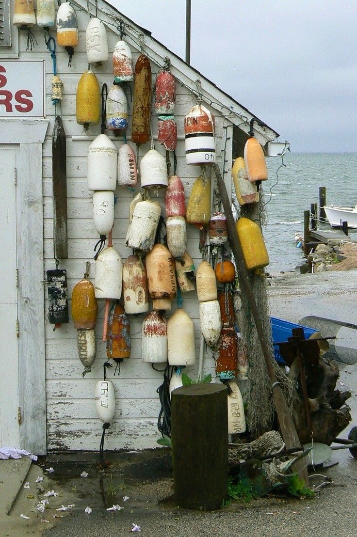 A weathered white seaside shack is adorned with a colorful assortment of old buoys hanging on the exterior wall. The buoys vary in shape, size, and color—mostly reds, yellows, and whites—adding a rustic, nautical charm. In the background, a cloudy sky looms over the ocean, with a dock and small boats nearby. The scene evokes a salty, coastal atmosphere full of character and maritime history.