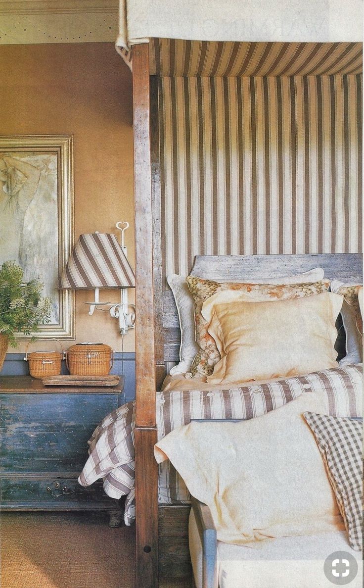 A cozy, vintage-inspired bedroom with a striped canopy bed featuring warm neutral bedding in florals and checks. A distressed wood nightstand sits beside the bed, topped with woven baskets and a small sconce with a matching striped lampshade. Earth-toned walls and an antique framed artwork complete the layered, rustic charm of the space.