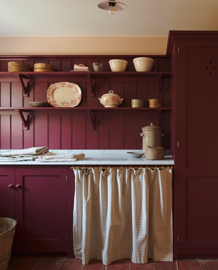 A moody traditional pantry with deep burgundy cabinetry and vertical tongue-and-groove paneling. The counter is styled with vintage red and white transferware, pottery, and folded linens. A gingham curtain hangs under the sink in place of cabinet doors.