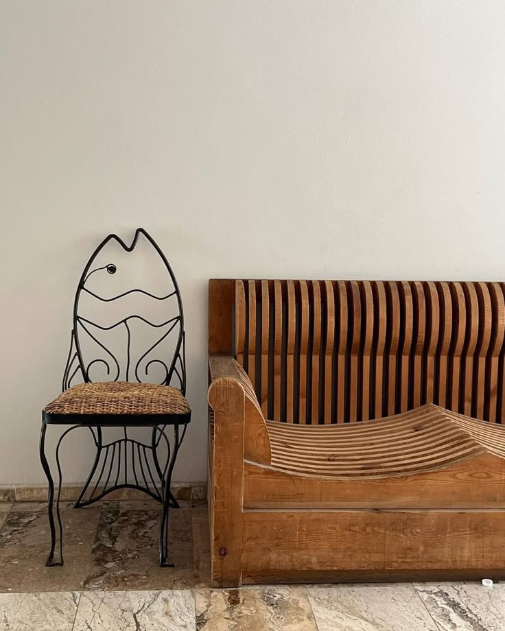 A minimalist seating arrangement against a plain white wall, featuring a whimsical black iron chair shaped like a fish with a woven rattan seat, next to a sculptural wooden bench with curved, slatted detailing and natural grain. The floor is made of mixed marble tiles, adding an earthy touch to the artistic and coastal-inspired space.