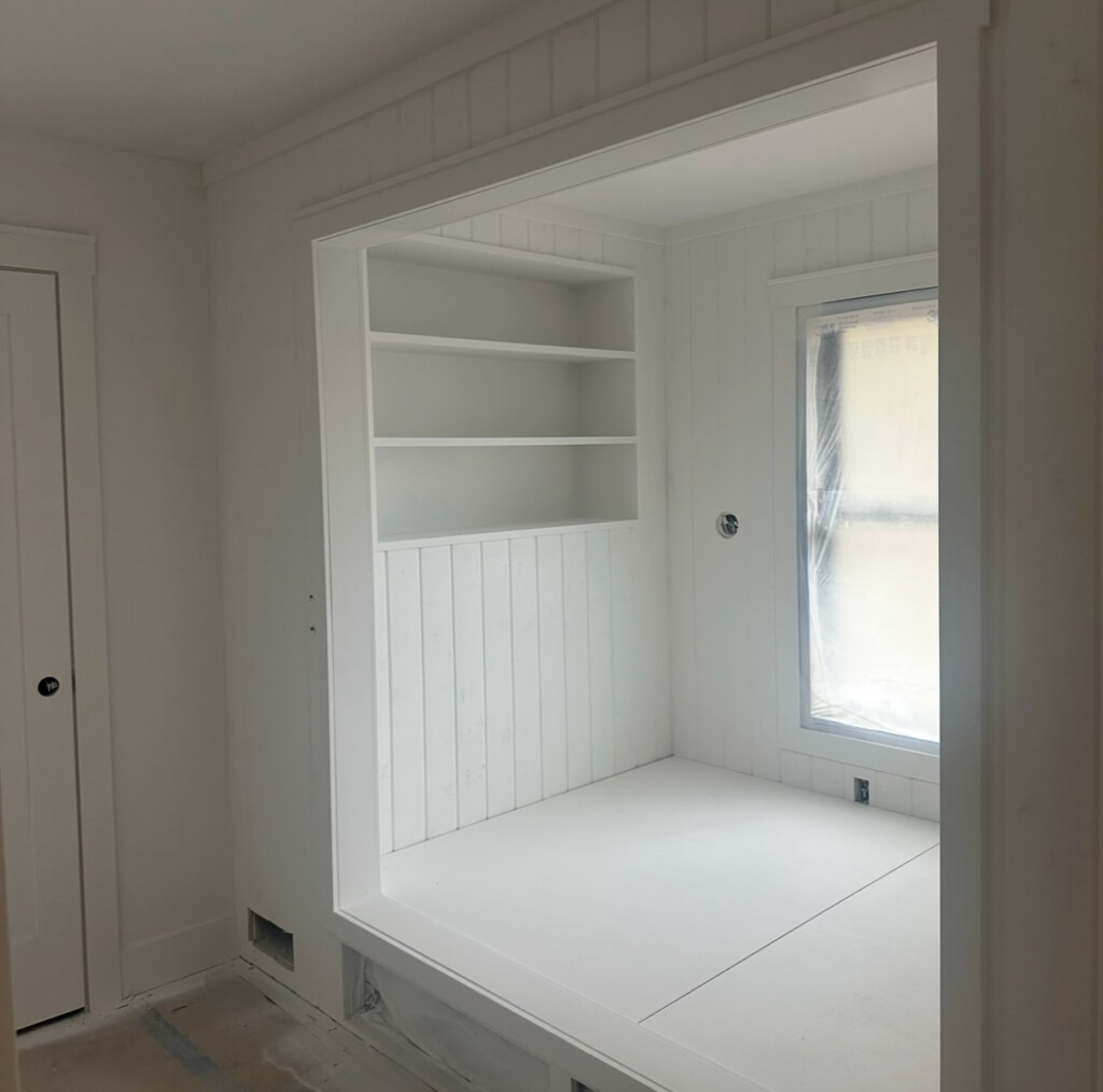 A cozy built-in bed nook in progress with vertical white shiplap, open shelving above, and a large window bringing in natural light.