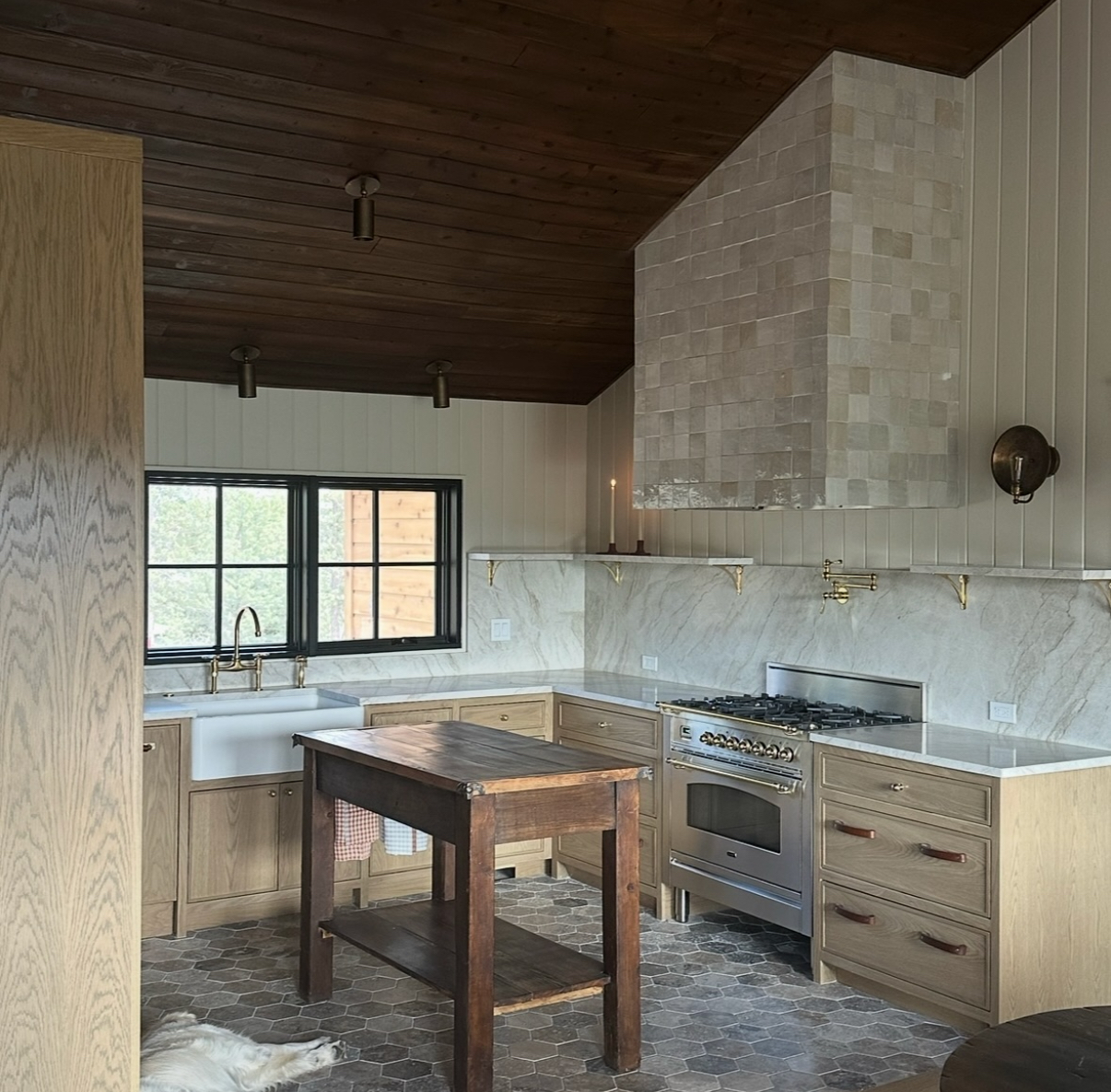 A warm, inviting kitchen featuring natural wood cabinetry, marble countertops, a Cle zellige tile hood, brass fixtures, and dark wood ceilings for contrast.