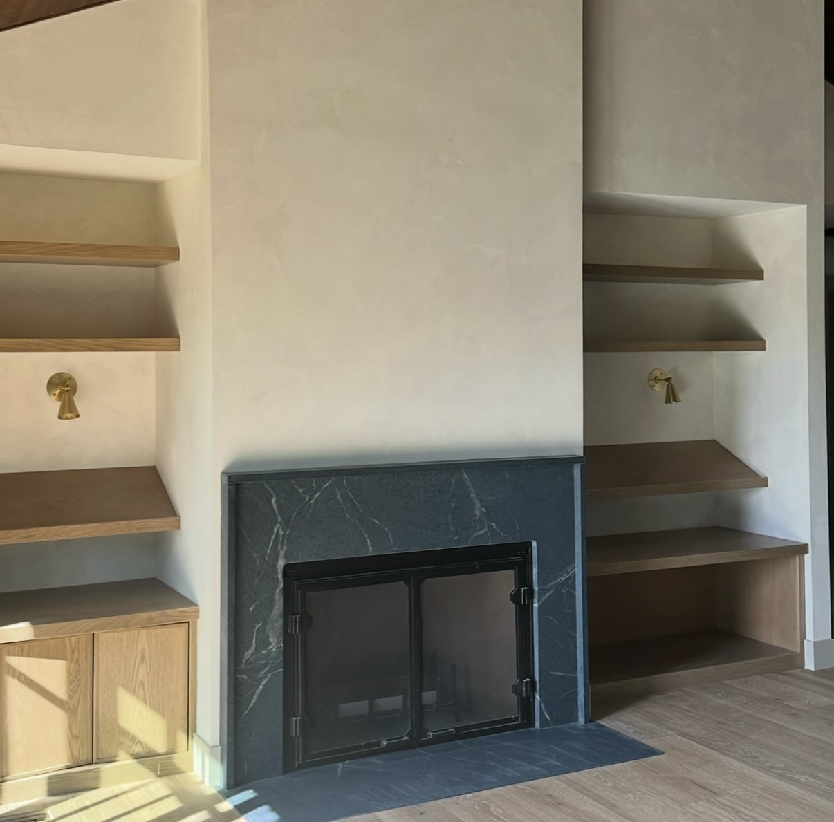 A modern fireplace with a deep stone surround, flanked by floating oak shelves and integrated sconces for layered, ambient lighting.