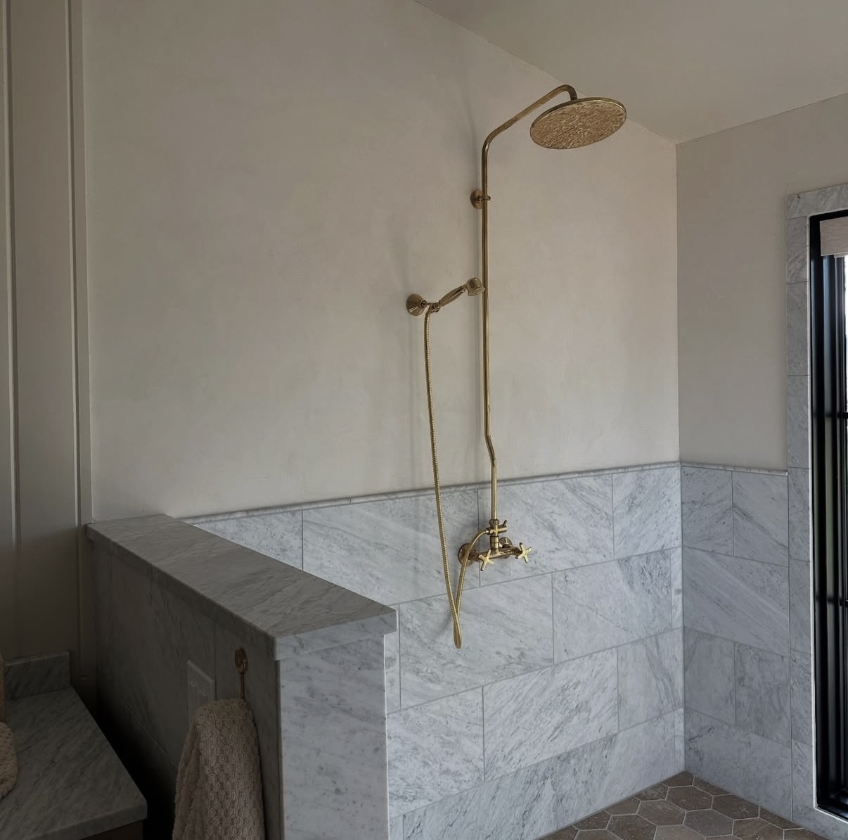 A spa-like shower with soft gray marble tile, a brass exposed shower fixture, and warm neutral walls creating an airy, serene retreat.