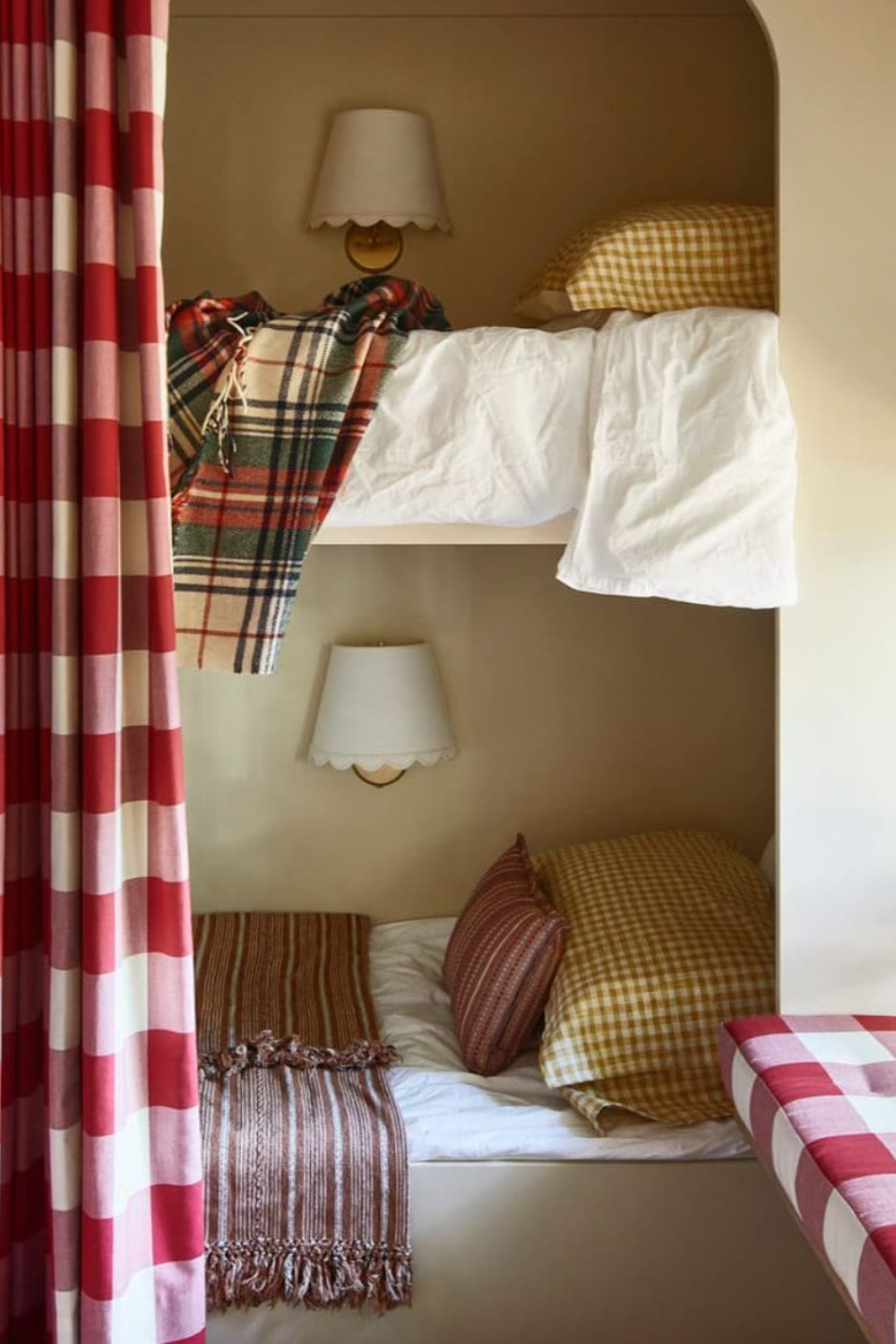 Built-in bunk beds with cozy autumnal bedding, including mustard gingham pillows, white sheets, and plaid blankets. A red and cream gingham curtain partially frames the scene, creating a private nook. Wall sconces with scalloped shades are mounted above each bed, and a striped brown throw blanket adds a rustic, layered touch. The overall palette feels warm, nostalgic, and perfectly suited for a cottage or cabin.