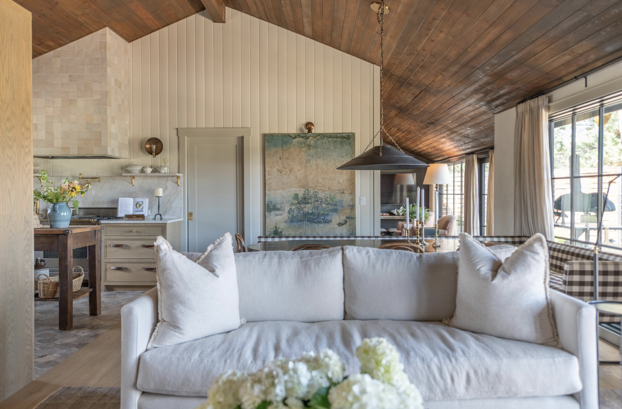 Open-concept living and kitchen space with warm, natural textures and a cozy, layered feel. A soft cream sofa with plush pillows sits in the foreground, facing a wood-paneled vaulted ceiling. The kitchen features a zellige tile hood, marble countertops, and open shelving with brass brackets. A moody vintage landscape painting anchors the dining area, lit by a large black pendant light. Neutral drapery, a checkered armchair, and floral arrangements add charm and softness throughout the space. Natural light pours in through large glass doors, framing a serene view of the outdoors.