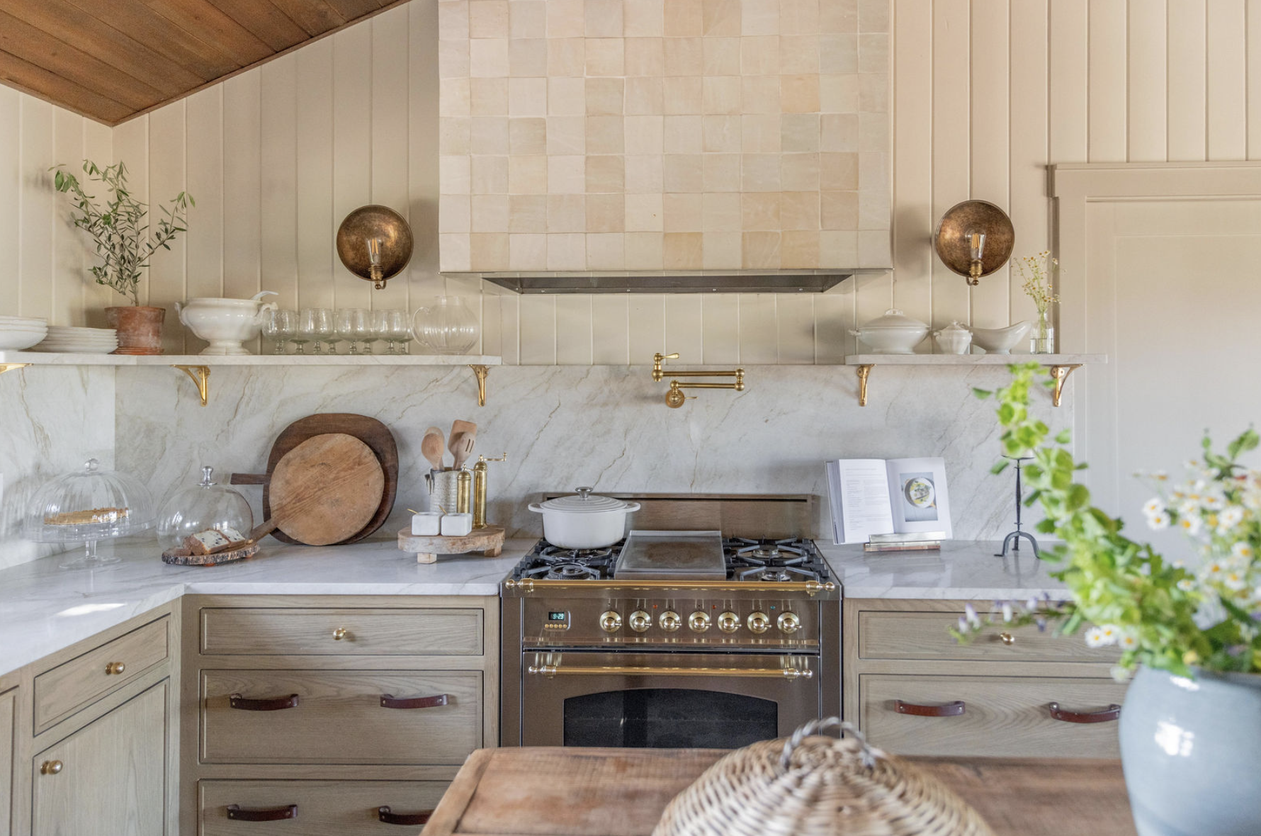 A warm and inviting kitchen with a rustic yet elevated design. The space features light wood cabinetry with brass knobs and leather pulls, a marble backsplash, and a creamy plastered range hood tiled in a subtle grid pattern. Open shelves hold simple white dishes and glassware, while brass accents—like a pot filler and sconce lights—add a touch of elegance. A mix of natural textures, including a woven basket, wooden cutting boards, and fresh greenery in a vase, brings the space to life with an organic, cozy feel.