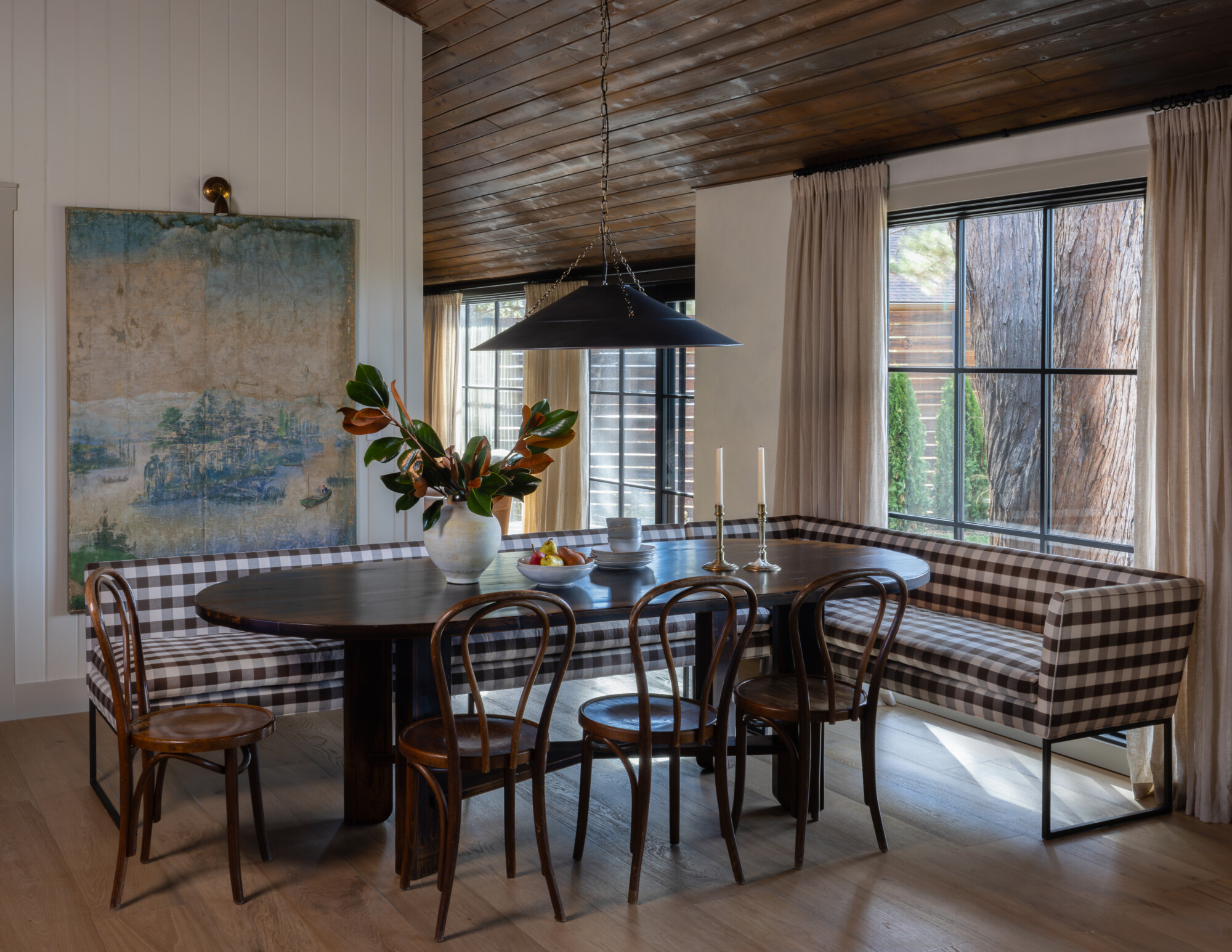 Cozy dining nook with a custom brown checkered banquette wrapping around a dark wood oval table. The space is styled with vintage bentwood chairs, linen curtains, and a statement black pendant light. A large antique-style landscape painting hangs on the paneled wall, and natural light pours in from black steel-framed windows, highlighting the warm wood ceiling and soft, layered textures. A ceramic vase with magnolia branches and a bowl of fruit bring life to the table, making the space feel lived-in and welcoming.