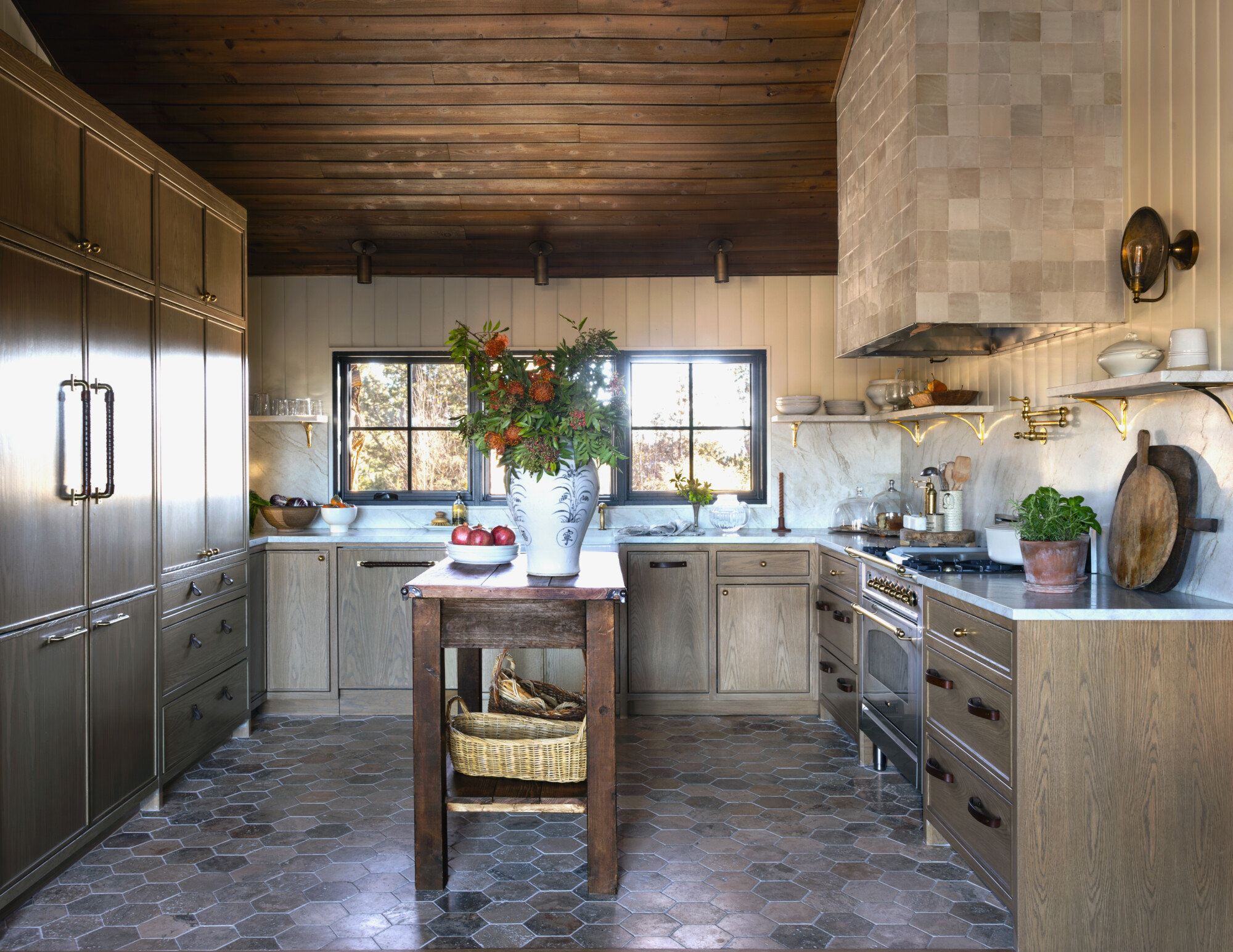 A warm, rustic kitchen with wood-paneled walls and ceiling, featuring a hexagon-tiled stone floor and natural wood cabinetry. A vintage-style range is framed by marble countertops, a handmade tile range hood, and open shelves with brass brackets. A wooden island holds a large vase with fresh greenery and pomegranates, adding a vibrant, lived-in feel. The space is bathed in soft natural light from two black-framed windows, highlighting its layered textures and inviting atmosphere.