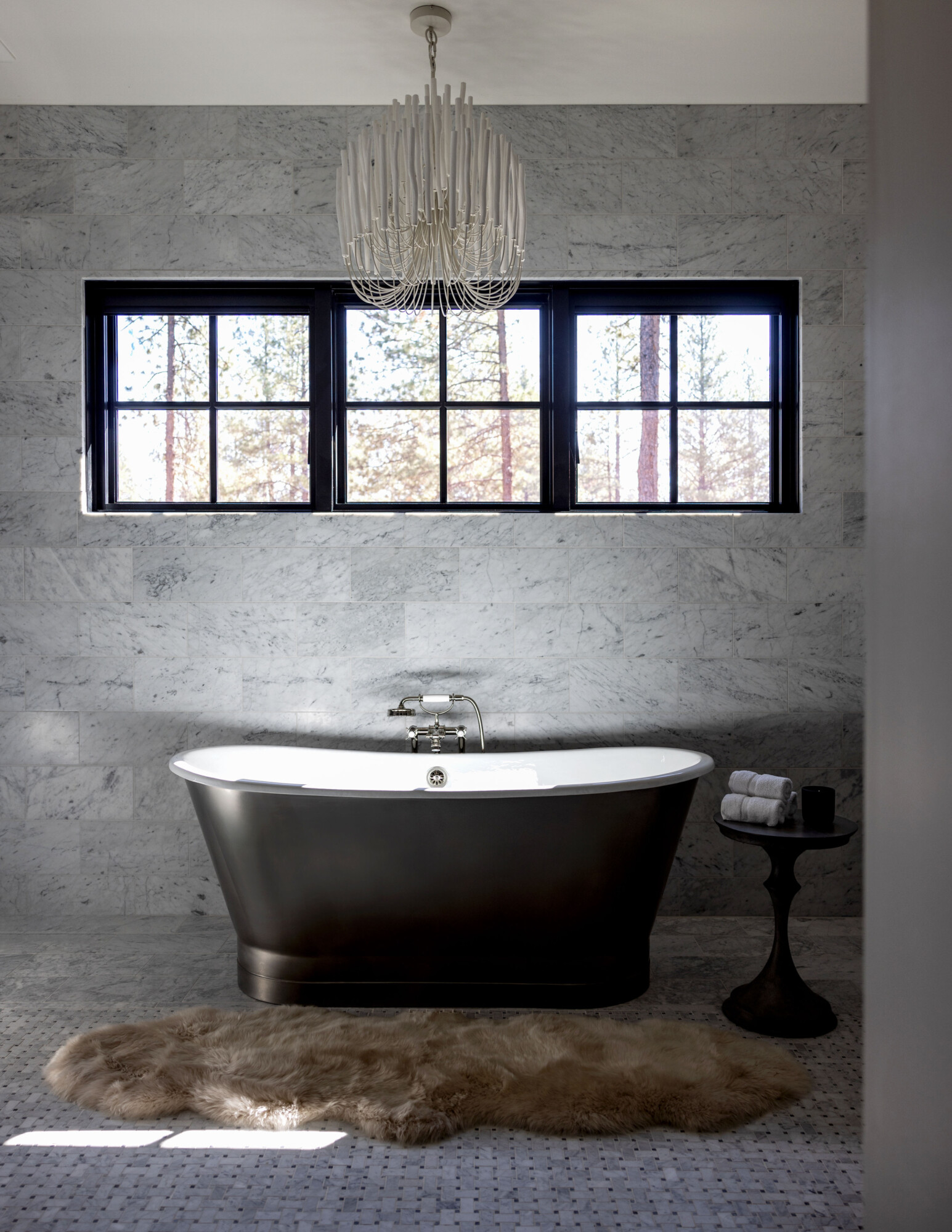 Sculptural freestanding soaking tub in a moody black finish, set against a backdrop of honed marble walls and mosaic tile floors. A plush taupe sheepskin rug adds warmth and softness underfoot, while a modern chandelier with cascading ceramic rods brings a touch of artful elegance above. Natural light pours through black-framed windows, casting soft shadows and enhancing the serene, spa-like feel of this minimalist primary bathroom. A black pedestal side table holds neatly rolled towels and a candle, completing the moment of calm luxury.