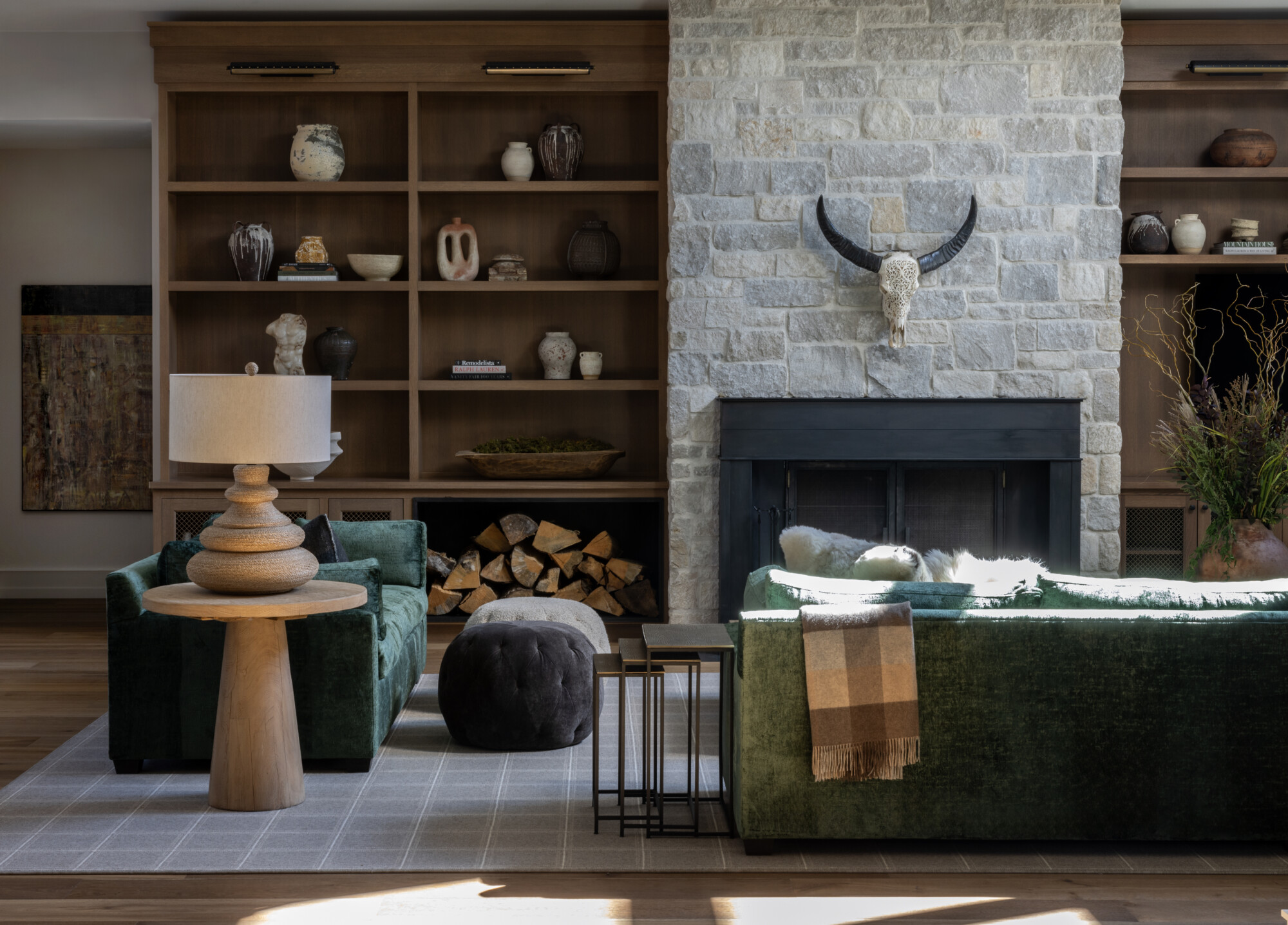 Cozy living room with rich textures and earthy tones. Two deep green velvet sofas face each other in front of a stone fireplace with a black metal surround and a sculptural horned skull mounted above. A built-in wood storage nook beneath the firebox adds rustic charm. Behind the seating area, natural oak built-ins are styled with ceramics, books, and collected objects. A sculptural stone table lamp rests on a chunky wood pedestal side table, paired with a dark tufted ottoman and nesting tables. A soft plaid throw drapes over the back of the sofa, completing this layered and grounded space