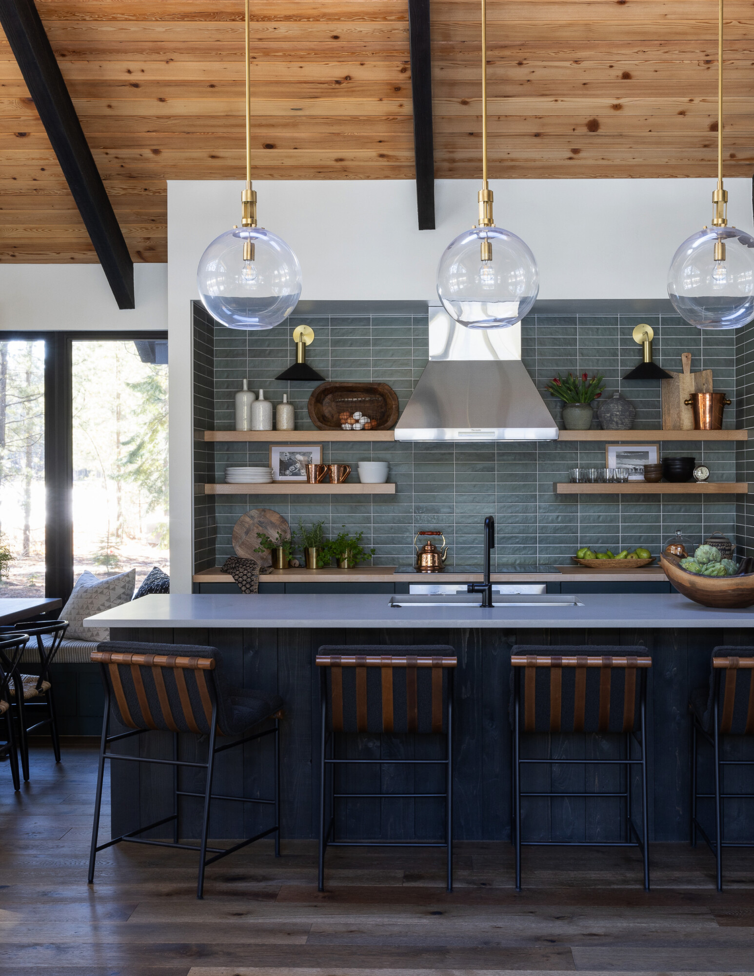 Modern mountain kitchen with vaulted wood ceilings, dark lower cabinetry, and a dramatic green tile backsplash. A white quartz island with integrated sink is lined with leather-strapped barstools, offering a warm contrast to the cool tones. Three oversized glass globe pendants hang overhead, adding a touch of glam. Open wood shelves display ceramics, copper accents, and greenery. The stainless steel hood and matte black sconces tie in modern elements, while the overall palette blends rustic charm with elevated design. Floor-to-ceiling windows bring in natural light and forest views.