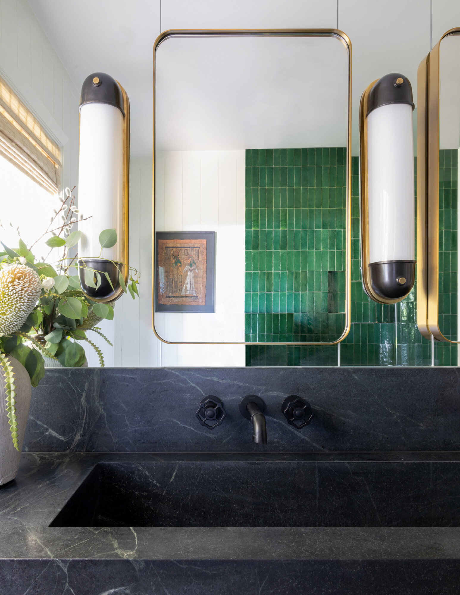 A striking bathroom vanity featuring a black marble integrated sink with matching backsplash and countertop. The faucet and knobs are matte black with a unique gear-like shape. Above the sink are brass-framed rectangular mirrors and contemporary sconces with black and white cylindrical shades. In the background, a wall of glossy green vertical tiles adds a vibrant pop of color, complemented by a neutral wood slat window shade and a potted floral arrangement to the left.