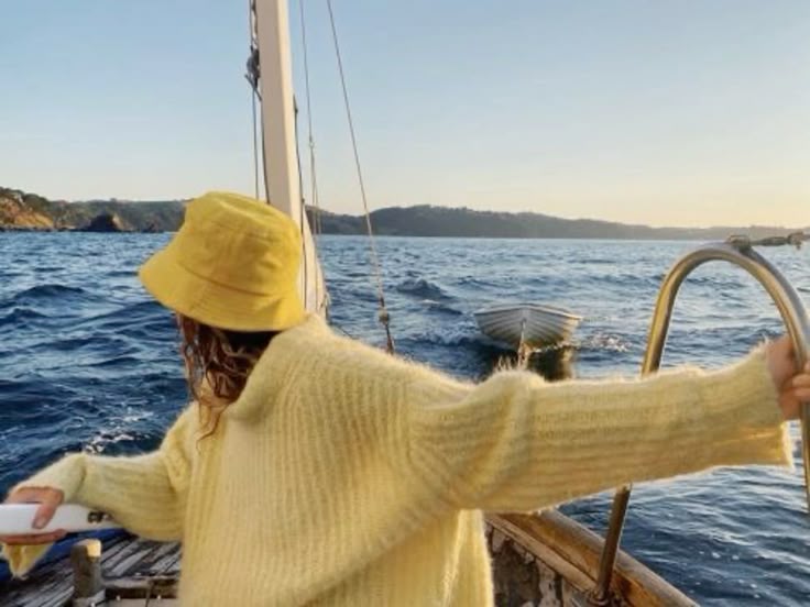 A woman in a soft yellow sweater and matching bucket hat stands at the edge of a sailboat, arms outstretched as the boat moves through deep blue ocean water. A small dinghy trails behind the sailboat, and the coastline is visible in the distance under a golden sunset sky. The moment feels carefree and peaceful.