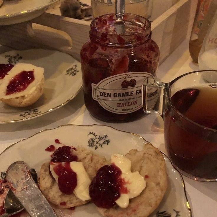 A cozy table setting with vintage floral china holding buttered scones topped with raspberry jam, a glass mug of tea, and a jar of Swedish raspberry jam labeled "Den Gamle Fabrik Hallon" with a spoon resting inside.