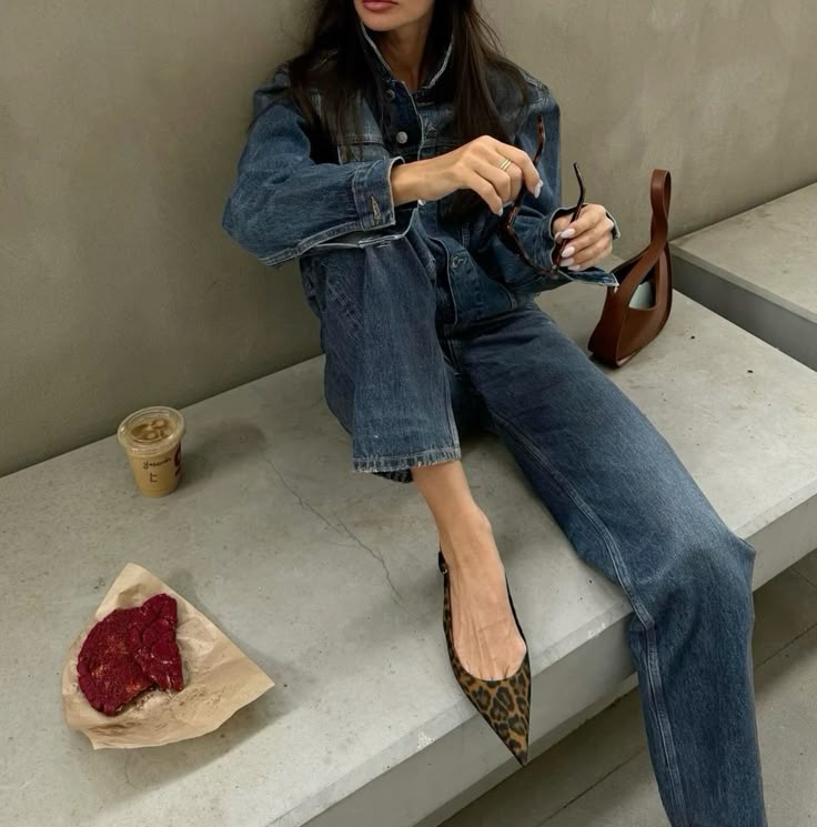 Woman sitting on a concrete bench wearing a denim-on-denim outfit with pointed leopard print heels, holding sunglasses in one hand. Beside her is an iced coffee, a large red velvet cookie on parchment paper, and a brown leather bag.