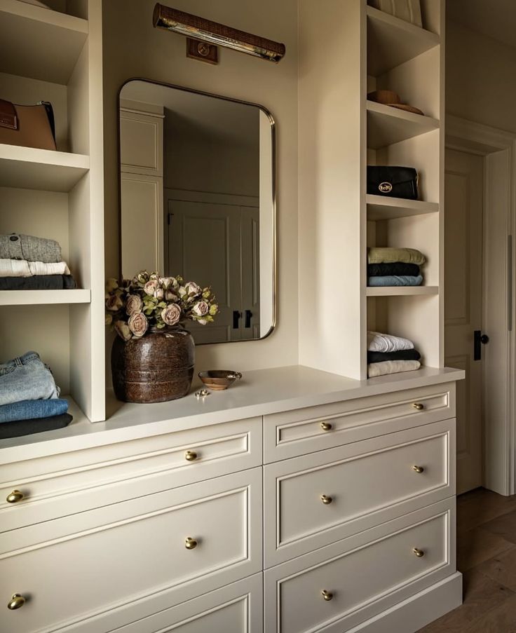 Built-in closet cabinetry in a warm neutral finish with brass knobs and open shelving. A central dresser-style counter is styled with a rustic vase of dried roses, a small dish, and a wall-mounted mirror with a brass picture light above.
