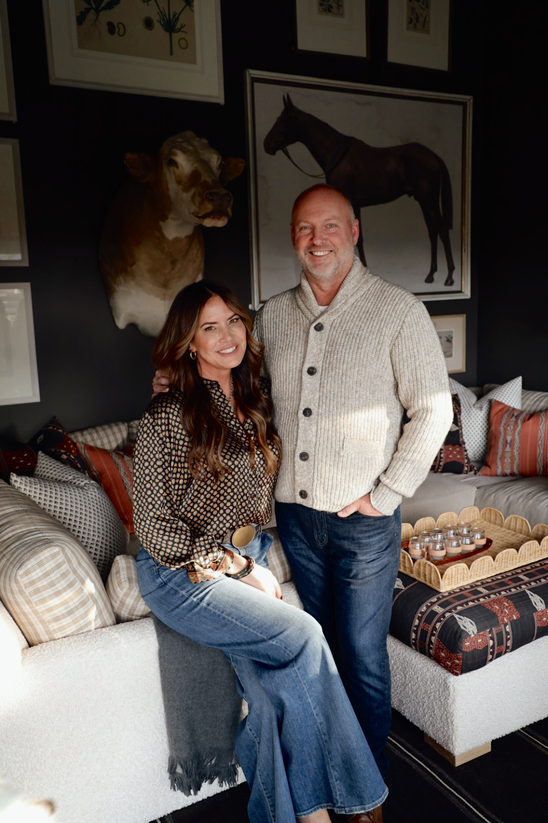 A smiling couple sitting together in a cozy, stylish living room with dark walls, patterned pillows, and framed artwork, including a horse portrait and mounted cow head. The woman is wearing a patterned blouse and wide-leg jeans, while the man is wearing a beige knit cardigan and jeans.