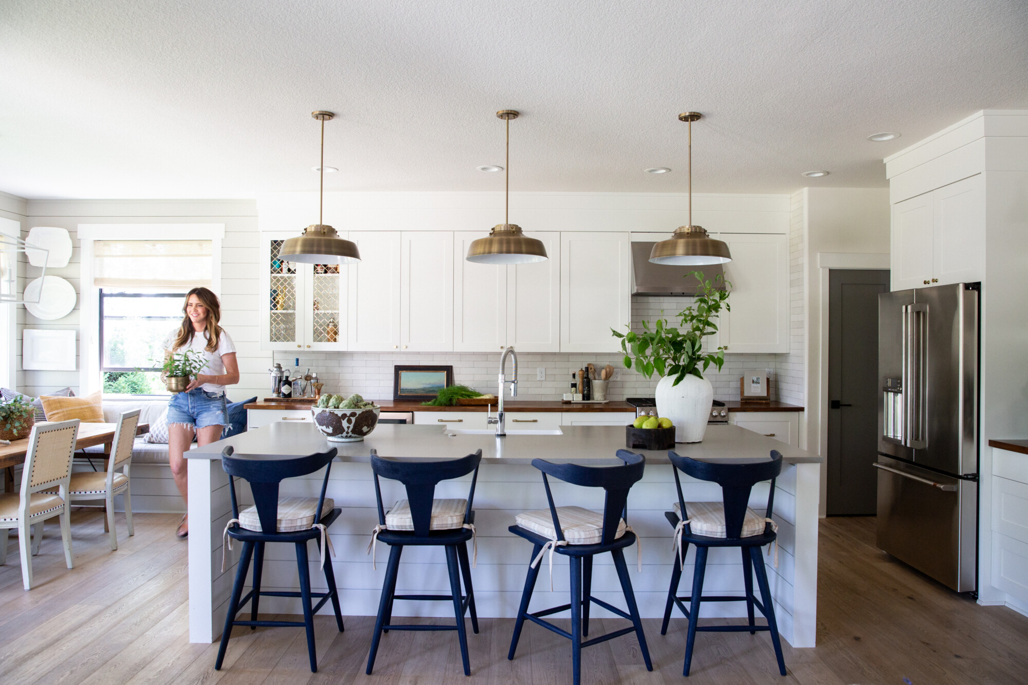 A bright and airy kitchen with white cabinetry, a large island, and three brass pendant lights. Four navy counter stools with plaid cushions line the island, which is styled with greenery and a bowl of fruit. A woman in denim shorts and a white tee carries a potted plant near the cozy dining nook, creating a casual and inviting atmosphere.