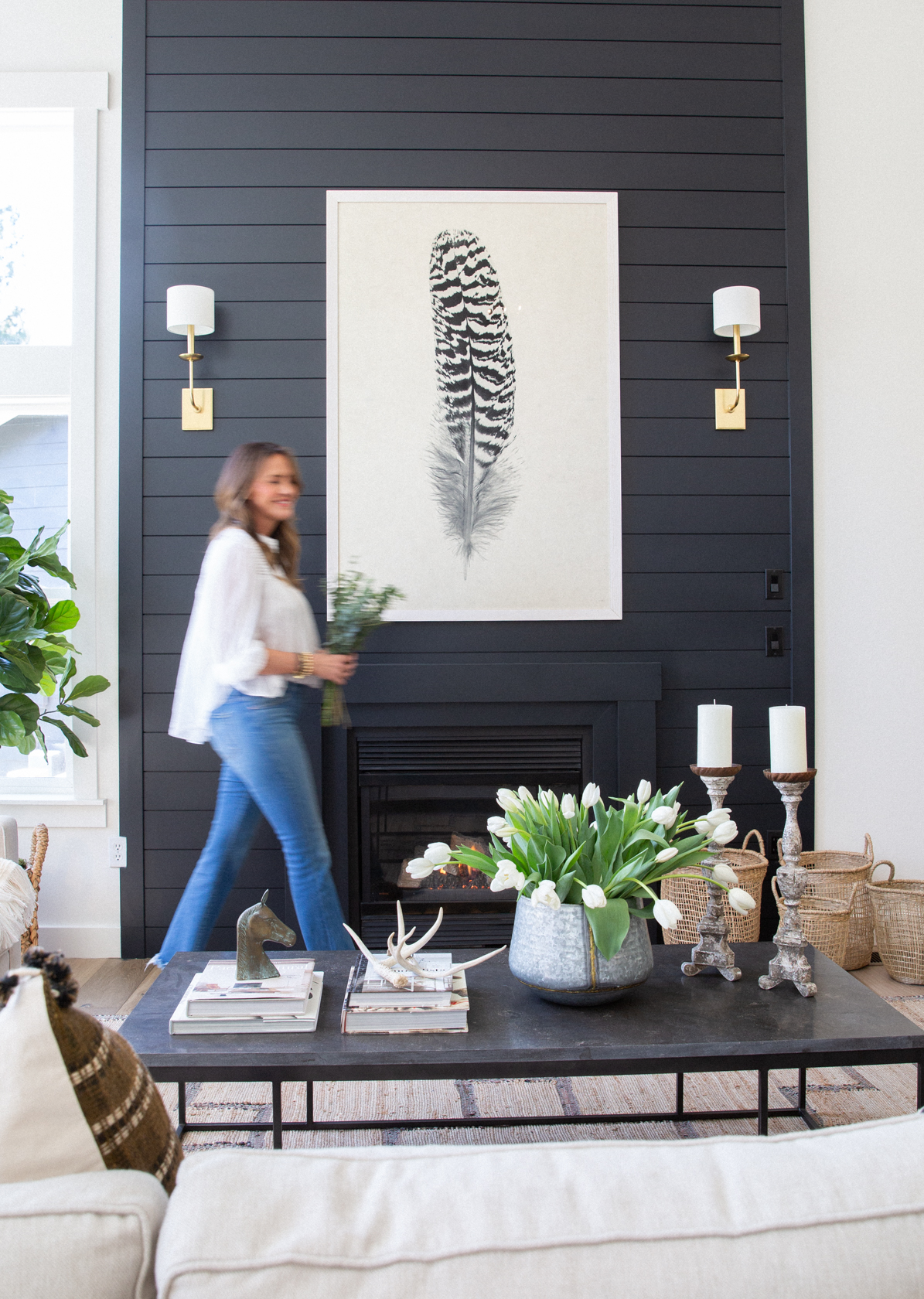 A modern living room with a black shiplap fireplace wall featuring a large feather artwork and brass sconces. A woman walks by holding greenery, while a black coffee table is styled with stacked books, antlers, candles, and a vase of fresh white tulips. Neutral sofas and woven baskets add warmth and texture to the cozy, layered space.