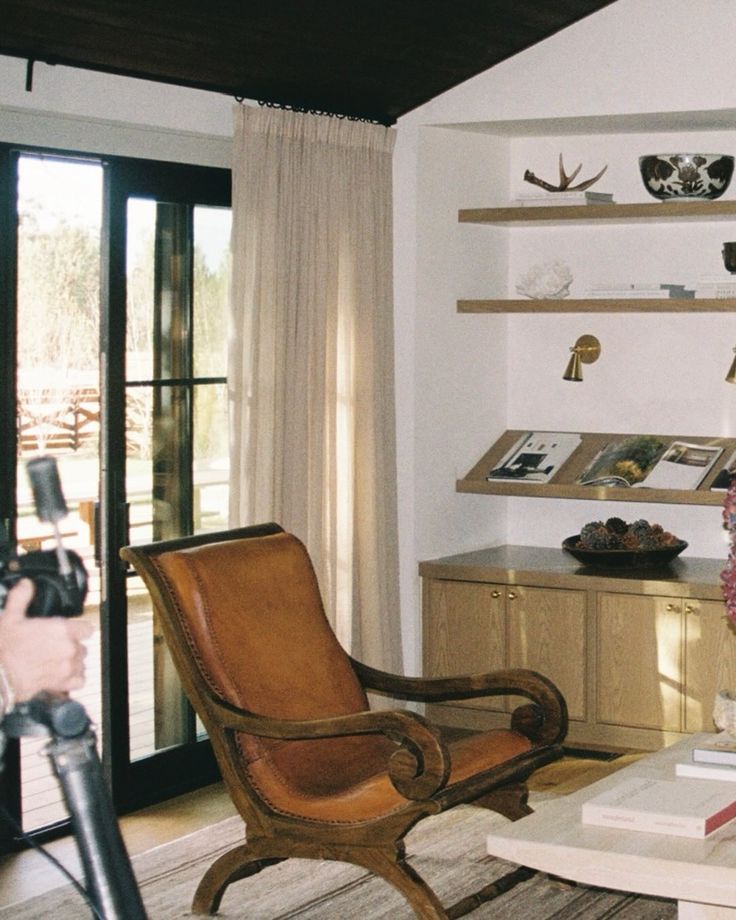 Warm library corner with leather sling chair, floating shelves, brass sconces, drapery.