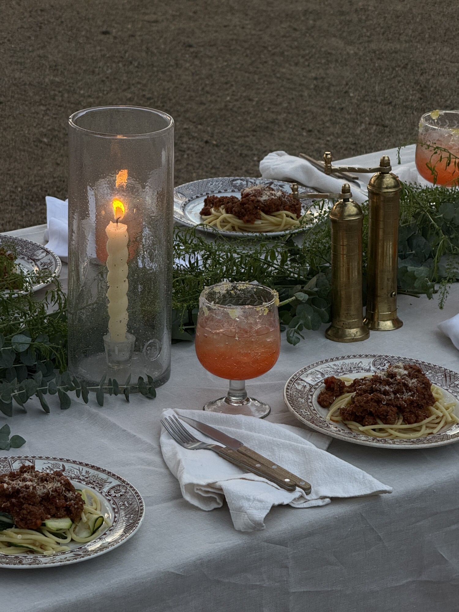 A rustic dinner table set with vintage patterned plates of pasta topped with ragu, Aperol sunset cocktails, a tall glass hurricane candle, and brass salt and pepper mills on a linen tablecloth with eucalyptus garland.